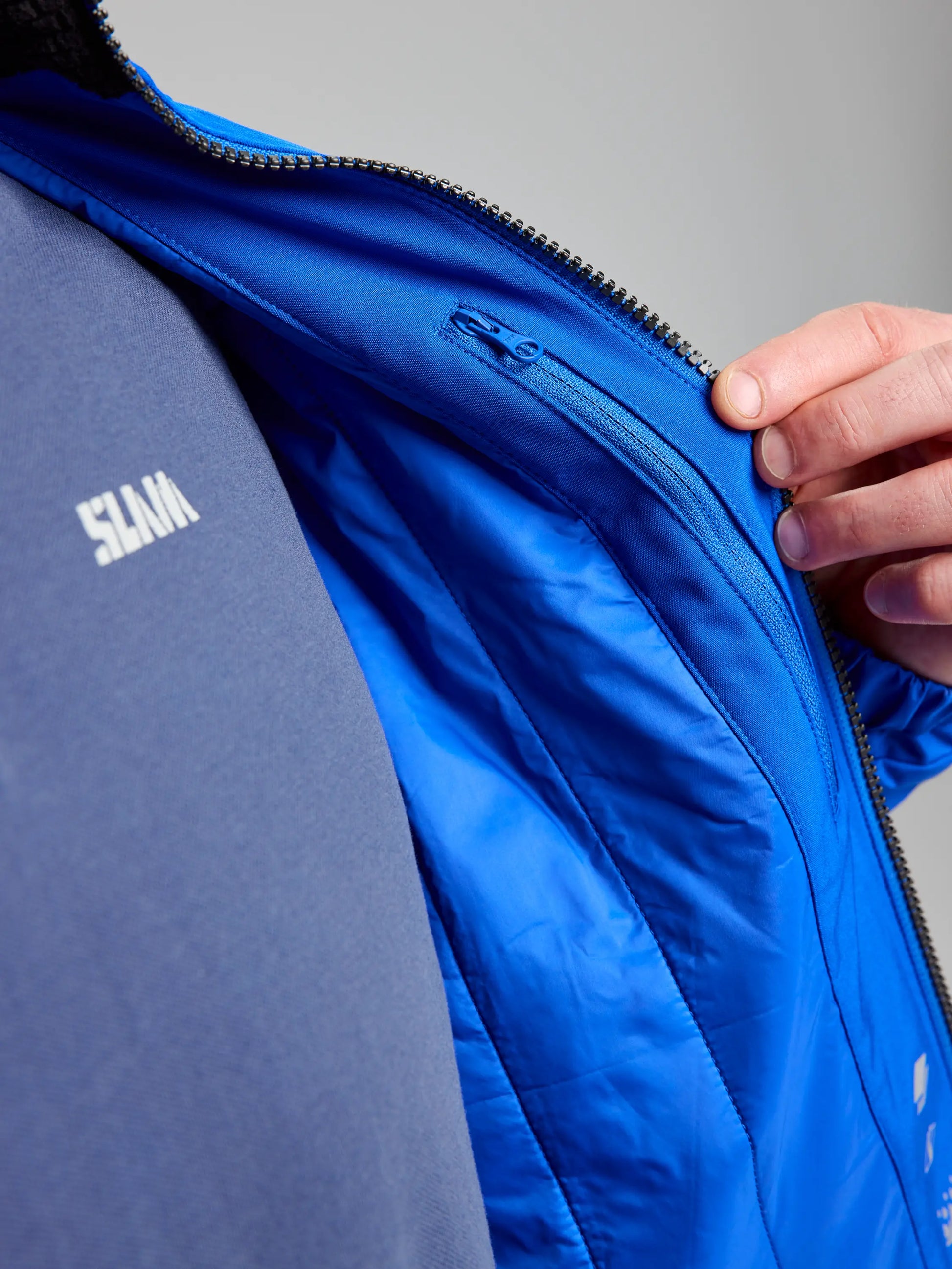 A person is unzipping the SLAM DOCK INSULATED JACKET CL in blue, revealing its sleek inner lining. The jacket's fabric looks shiny and smooth, with a navy blue shirt that has white text underneath. A hand grips the zipper pull against a plain light gray background.