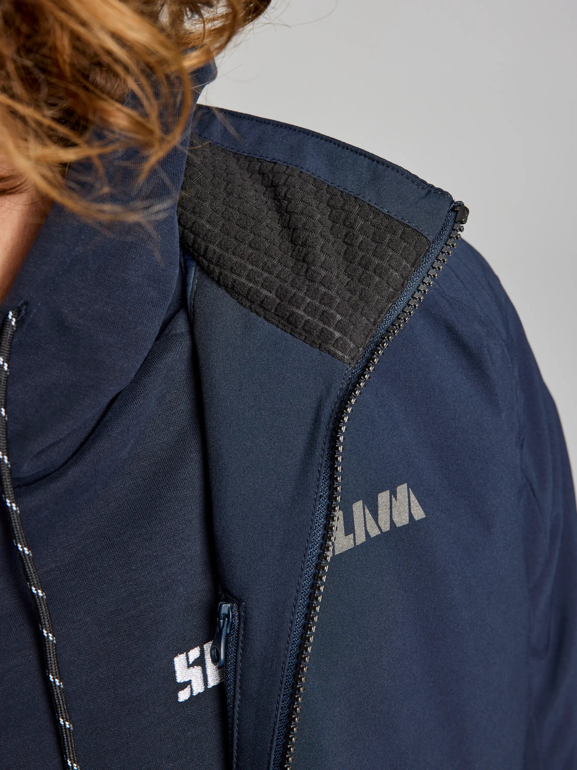 Close-up of a person wearing the DOCK INSULATED JACKET CL from SLAM, featuring a navy color with a partially visible silver logo on the chest. The jacket showcases a textured black shoulder patch and waterproof material, along with an exposed zipper and fleece-lined collar. Part of the person's hair is visible on the left.