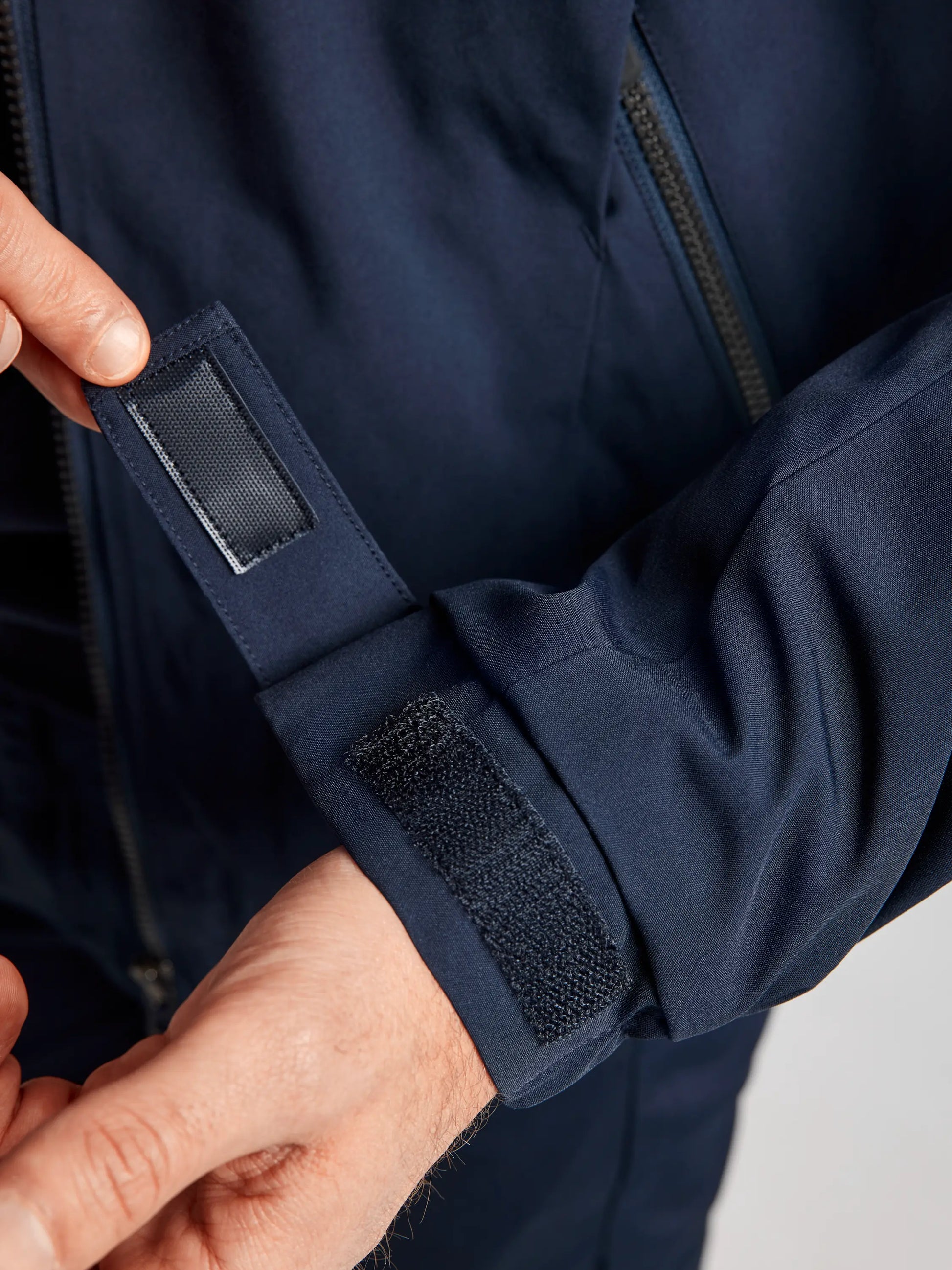 A person adjusts the sleeve of the DOCK INSULATED JACKET CL by SLAM in navy blue, tightening it with a Velcro strap. The focus is on their hands and the sleeve's detailing, with a fleece-lined collar and partially visible zipper on this waterproof jacket.