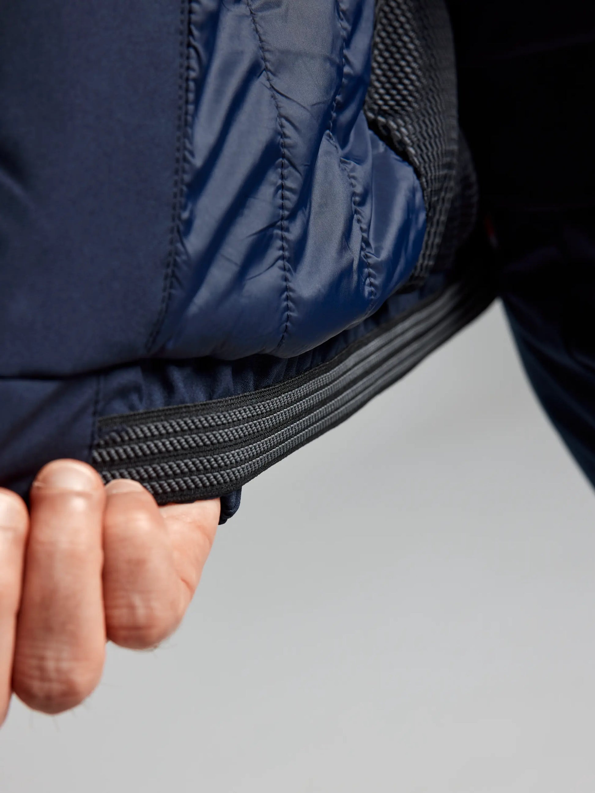 A person adjusts the bottom elastic cord of the DOCK INSULATED JACKET CL, a men's nautical winter jacket from SLAM, featuring a navy blue quilted design with smooth fabric and visible stitching. The fleece-lined collar promises extra warmth as the hand grasps the cord against a neutral background.