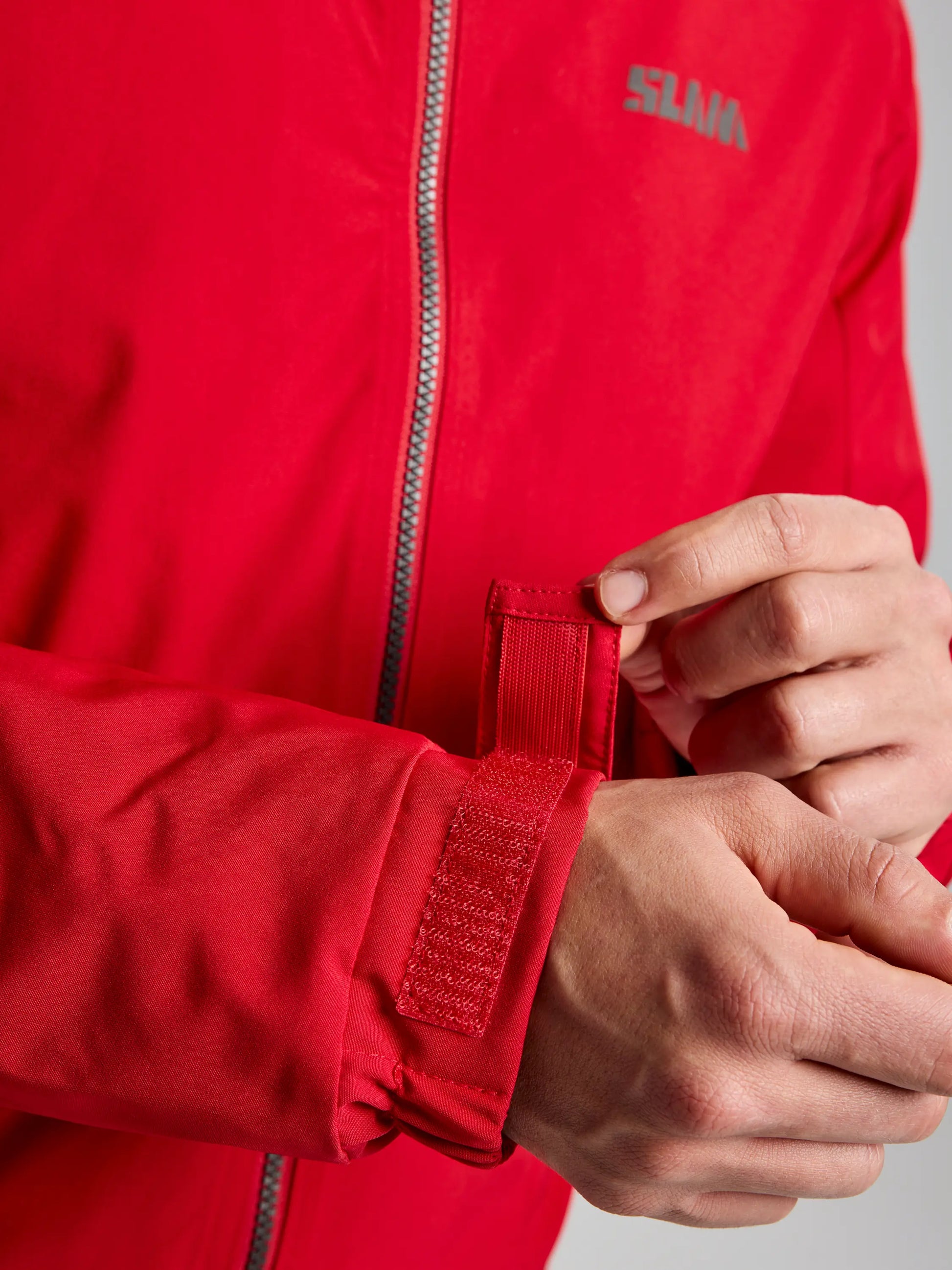 Someone is adjusting the Velcro strap on the cuff of a red DOCK INSULATED JACKET CL, a men's nautical winter jacket by SLAM. The jacket features a visible zipper and a small logo on the chest, along with a fleece-lined collar for added warmth. The focus is on the hands and jacket, set against a neutral background.