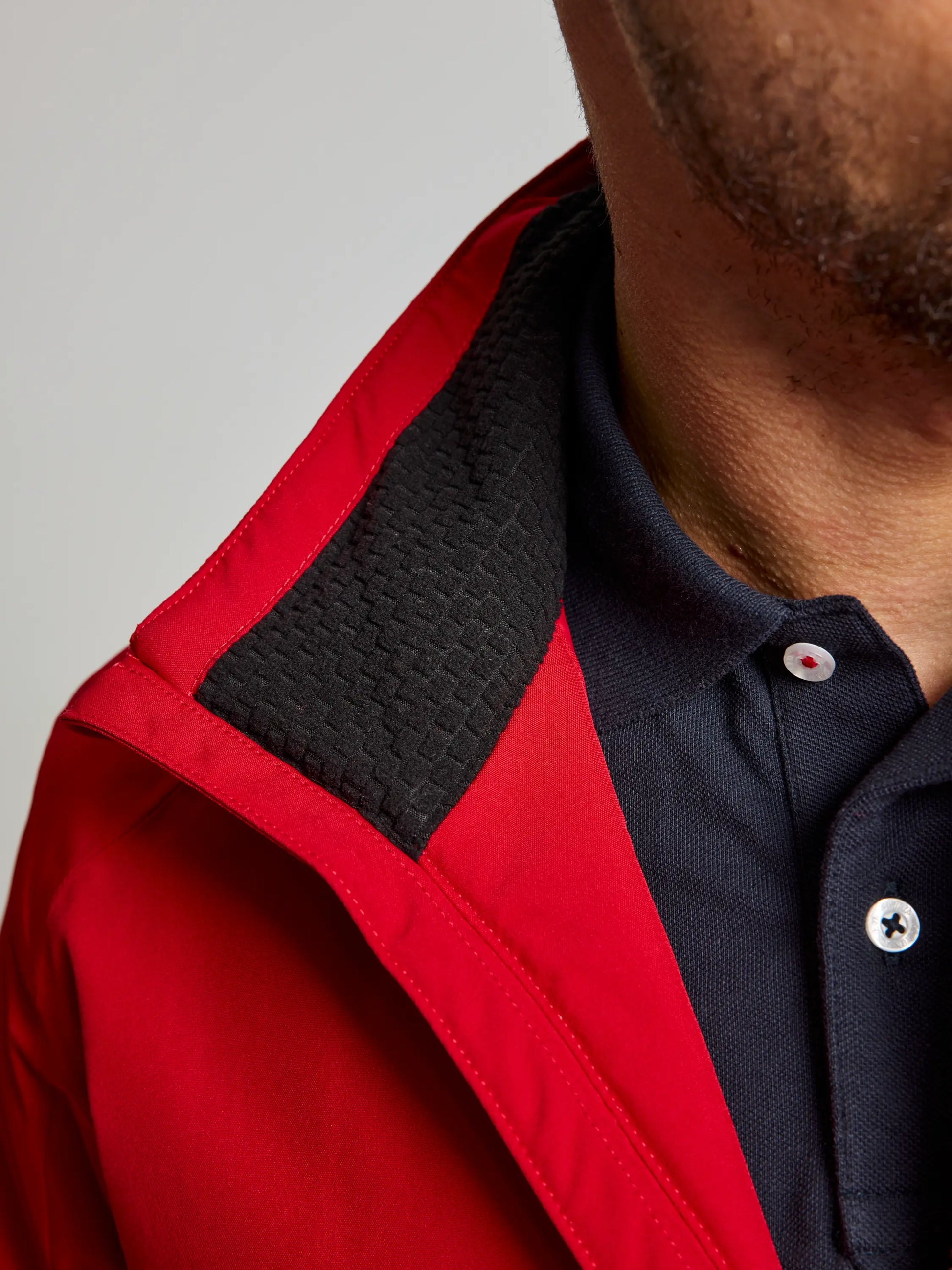 A detailed view showcases a person dressed in the DOCK INSULATED JACKET CL by SLAM, a red men's nautical winter jacket. It highlights the black textured, fleece-lined collar with a navy blue collared shirt worn underneath. The focus remains on the clothing details, capturing the top of their shoulder and a portion of their face.