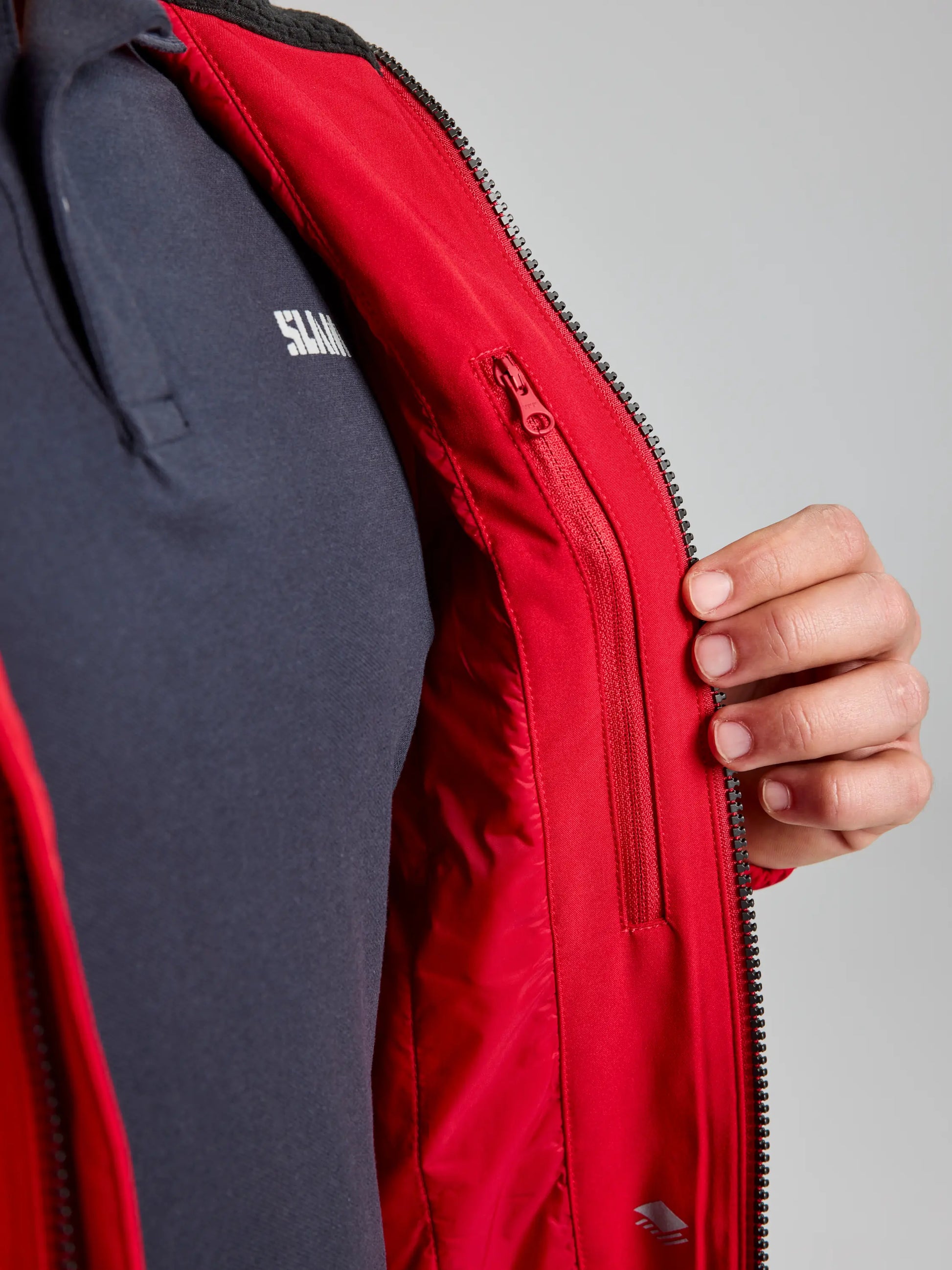 A person displaying the inside pocket with a zipper of a DOCK INSULATED JACKET CL by SLAM, showcasing its inner design, fleece-lined collar, and pocket detail. The individual is dressed in a dark blue polo shirt featuring white text on the chest.