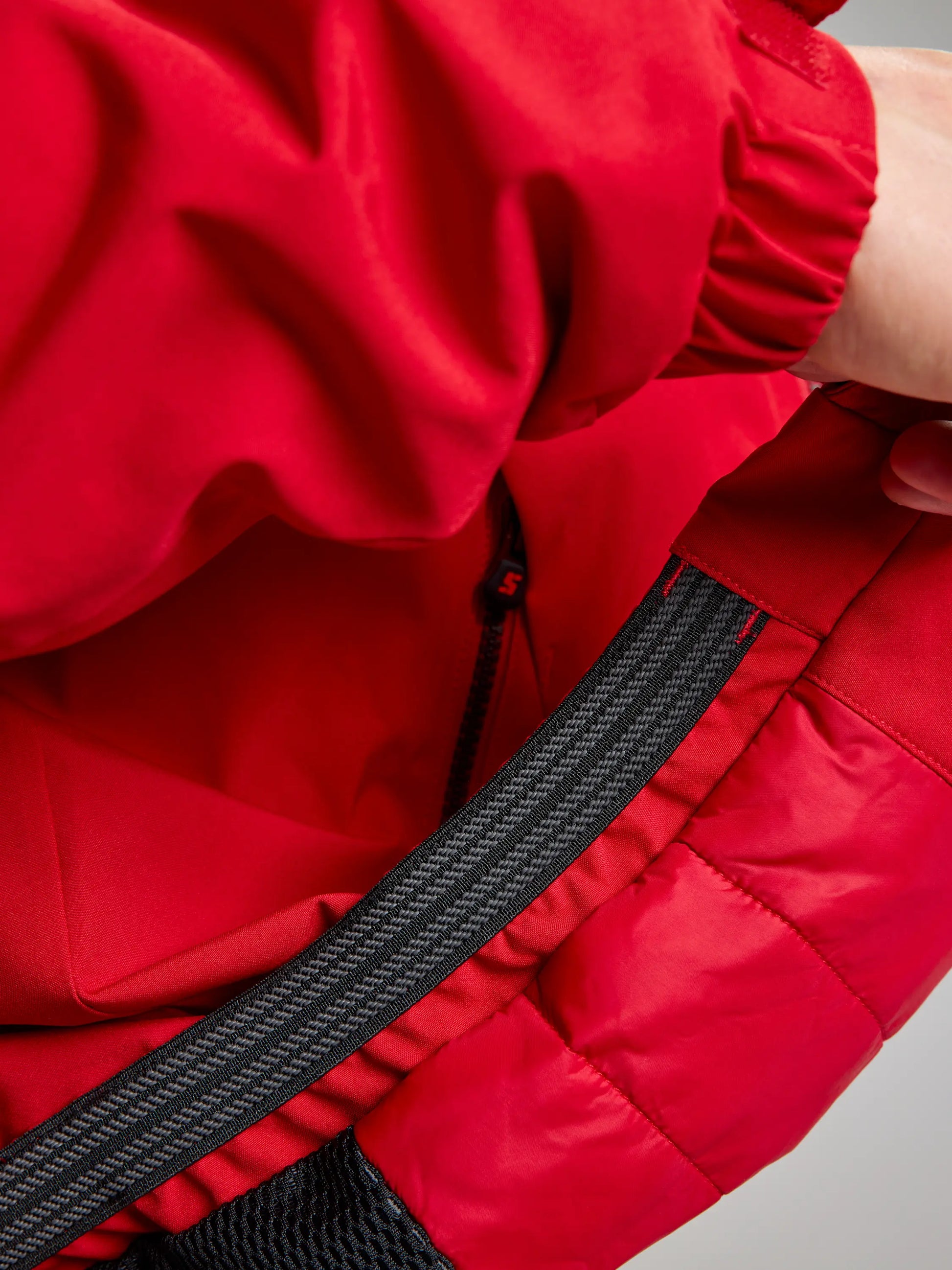 Close-up of someone wearing the SLAM DOCK INSULATED JACKET CL in red, showcasing its partially open zipper. The jacket includes an elastic band, textured lining, and a fleece-lined collar. The individual's hand can be seen pulling on the zipper.