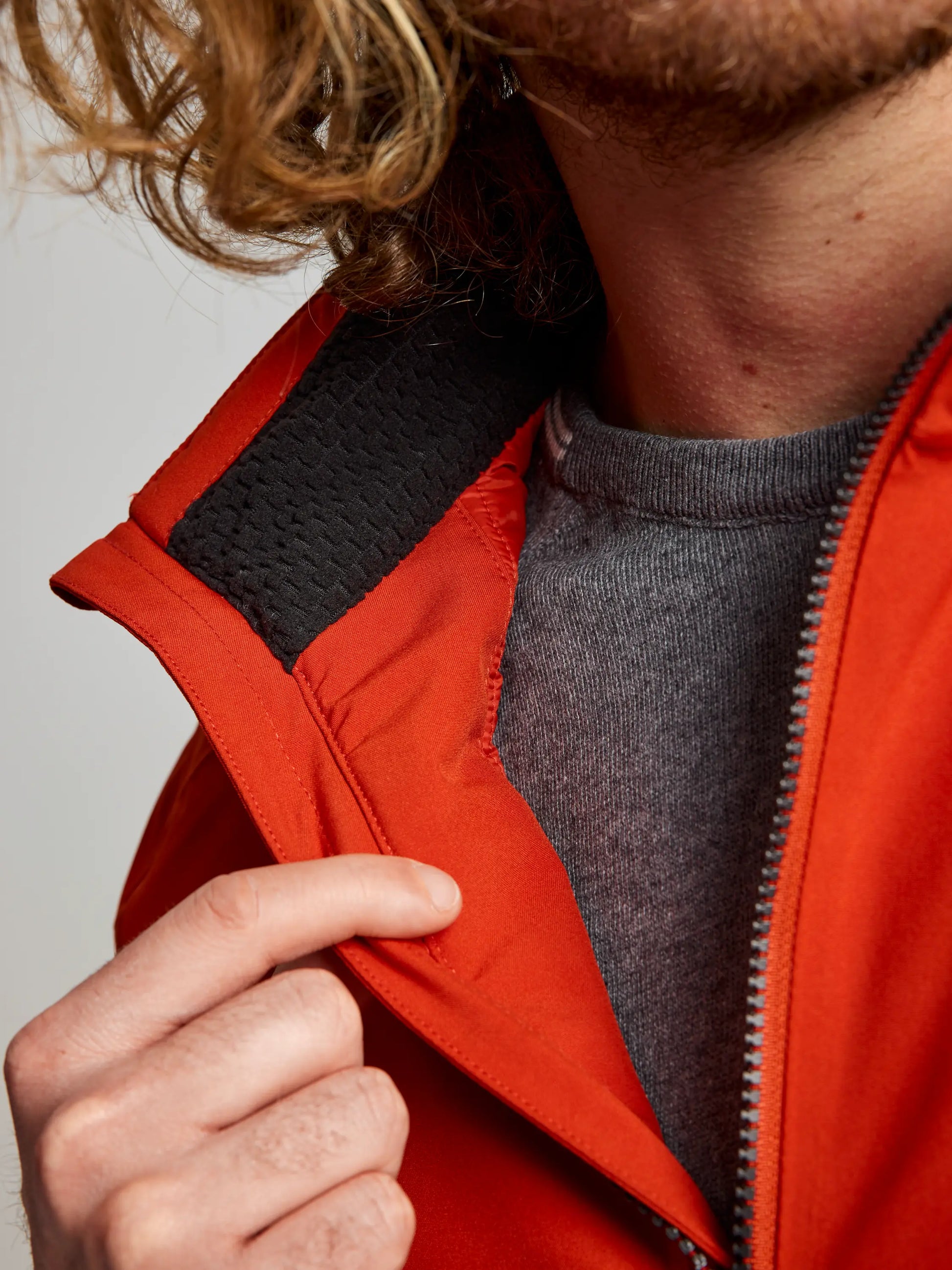 A close-up shows someone wearing the SLAM DOCK INSULATED JACKET CL, a men's nautical winter jacket in orange with a gray fleece-lined collar, layered over a gray shirt. They are holding the edge of the collar with their right hand, displaying its textured lining. The individual has shoulder-length curly hair.