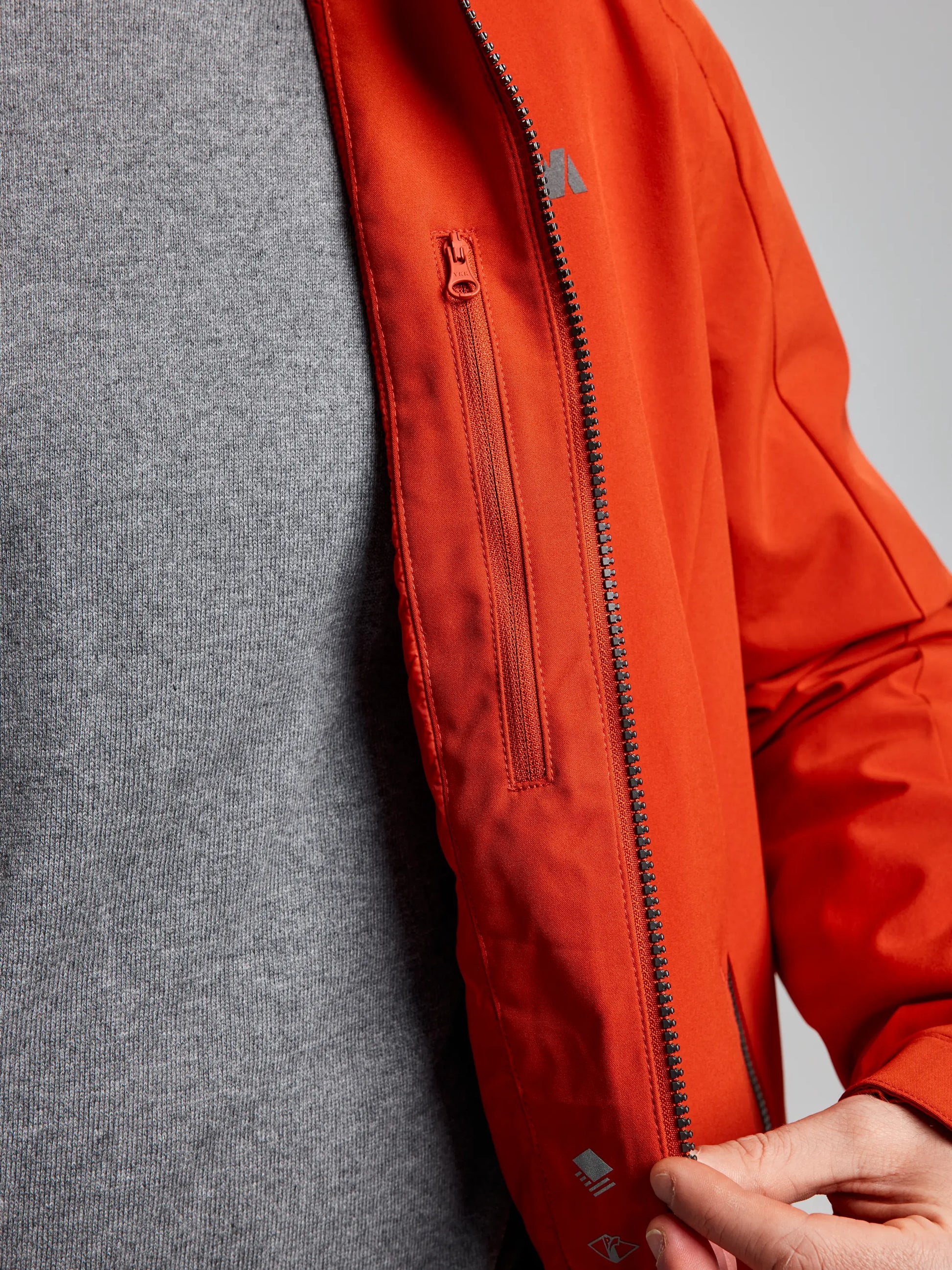 Close-up of a person wearing the DOCK INSULATED JACKET CL by SLAM over a gray shirt. The sleek, waterproof design showcases visible stitching and is partially unzipped, revealing the zipper. The hand adjusts the fleece-lined collar, adding warmth and style in its red-orange hue.