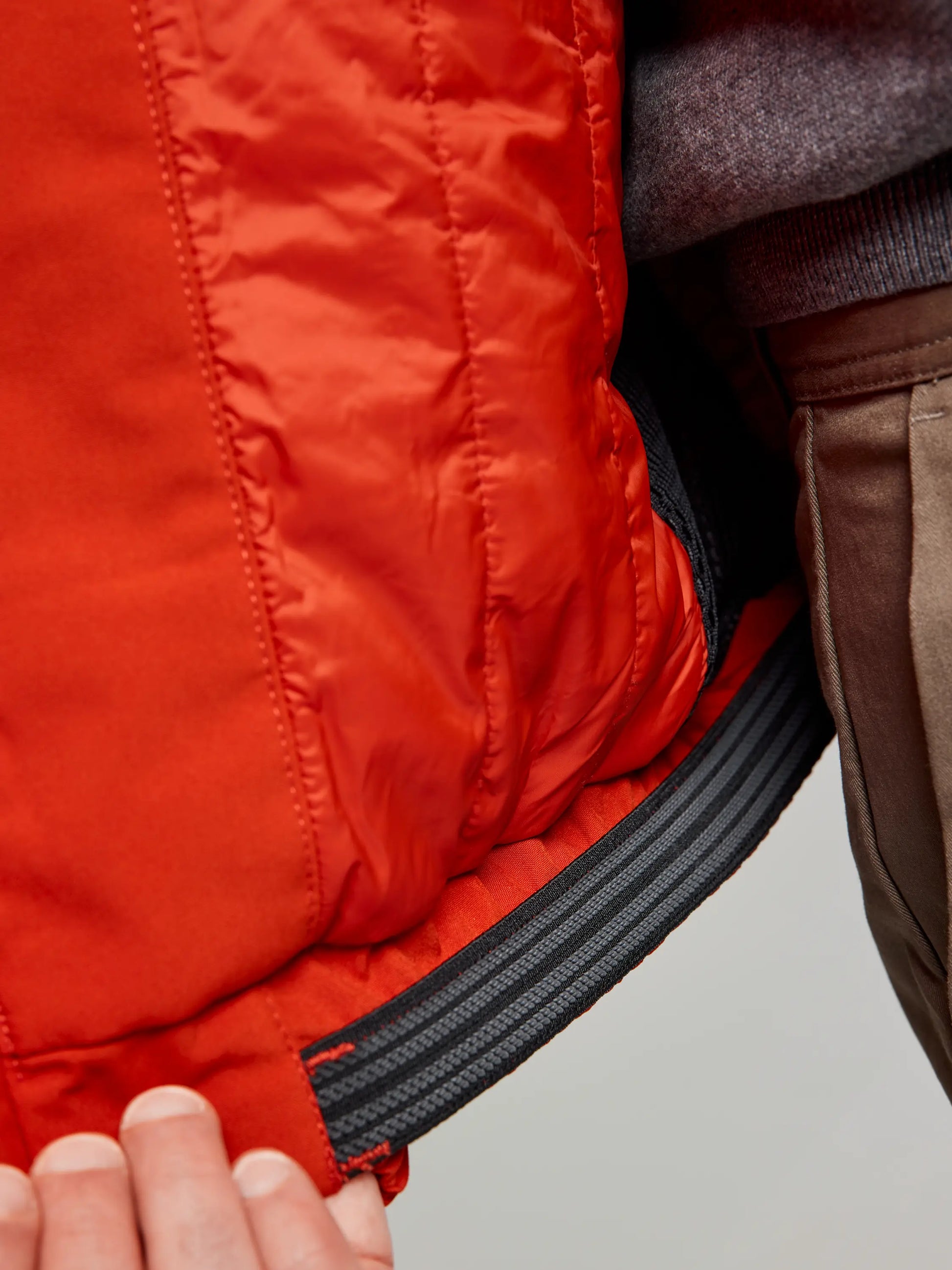 Close-up of a person holding the interior lining of a red SLAM DOCK INSULATED JACKET CL, revealing a black, elasticized waistband. A fleece-lined collar peeks out over their gray garment underneath.