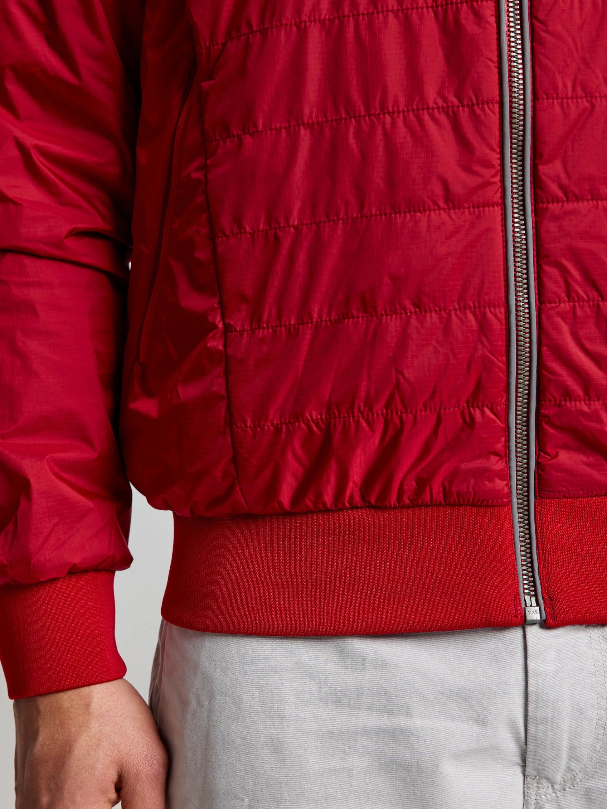 The image highlights an individual wearing the SLAM ACTIVE GRAPHENE SHORT JACKET CL, featuring vibrant red polyester wadding and a noticeable silver zipper. The jacket's ribbed cuffs are paired with light gray pants, drawing attention to the fabric texture and stitching details.