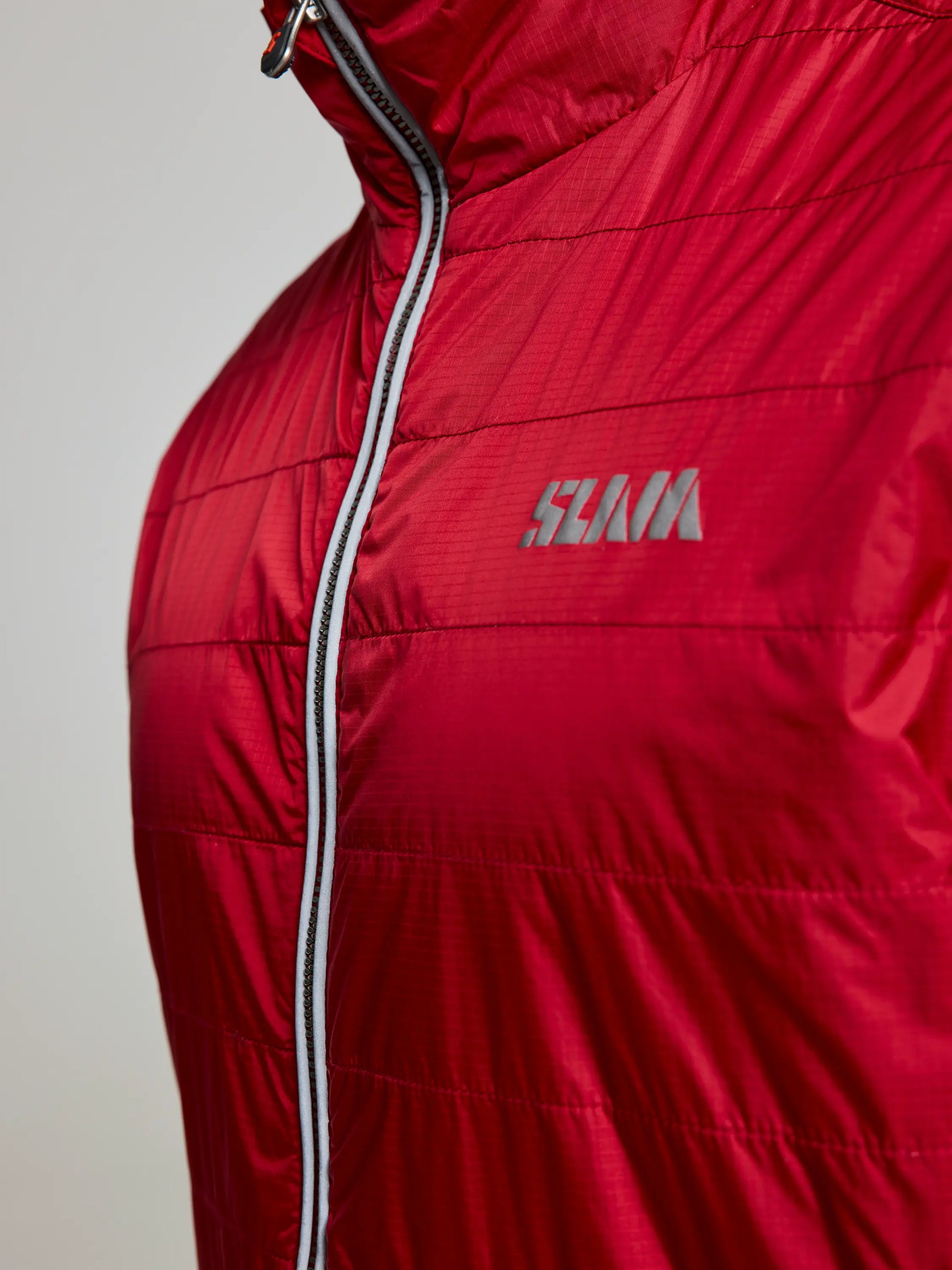 Close-up of the SLAM ACTIVE GRAPHENE SHORT JACKET CL, showcasing its red hue with a vertical zipper and white piping. Made from recycled nylon ripstop, this men's jacket features a stitched pattern and a distinctive logo on the left side of the chest.