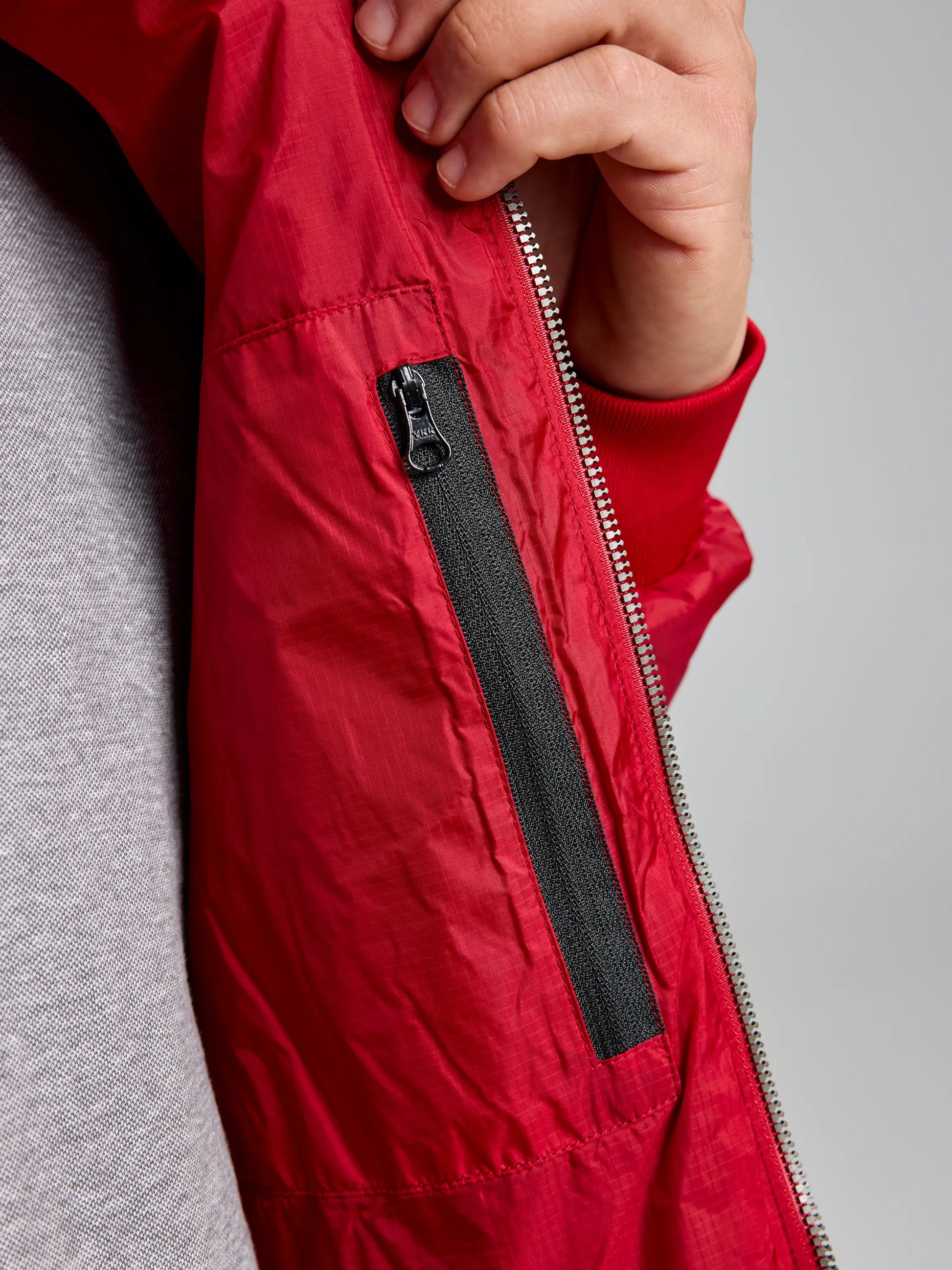 A person is holding open a red ACTIVE GRAPHENE SHORT JACKET CL by SLAM, showcasing a vertical black zippered pocket on the inner lining. Crafted with recycled Nylon Ripstop, the jacket is partially unzipped, revealing the gray shirt worn underneath. The hand rests close to the zipper.