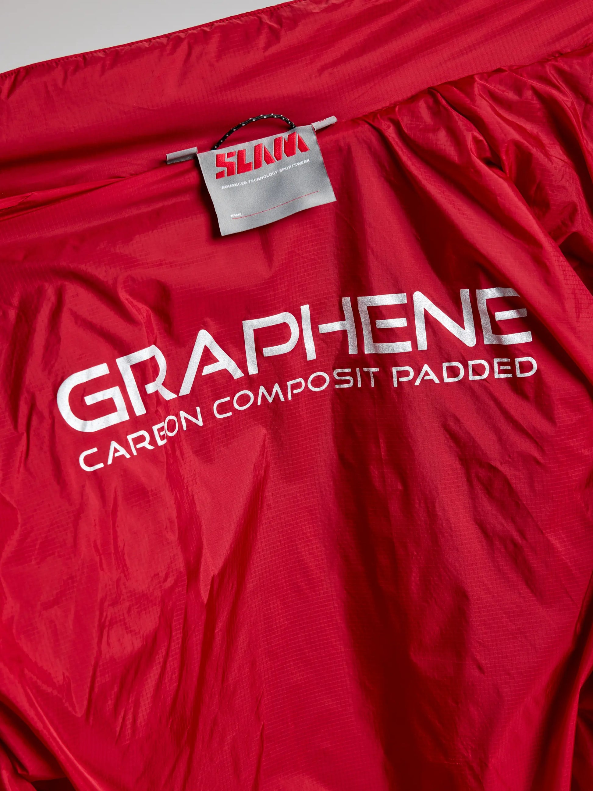 This men's jacket, branded as SLAM's "ACTIVE GRAPHENE SHORT JACKET CL," features a red fabric with a label indicating "SLIM" and prominently displays the text "GRAPHENE CARBON COMPOSITE PADDED." It is constructed from recycled nylon ripstop material.