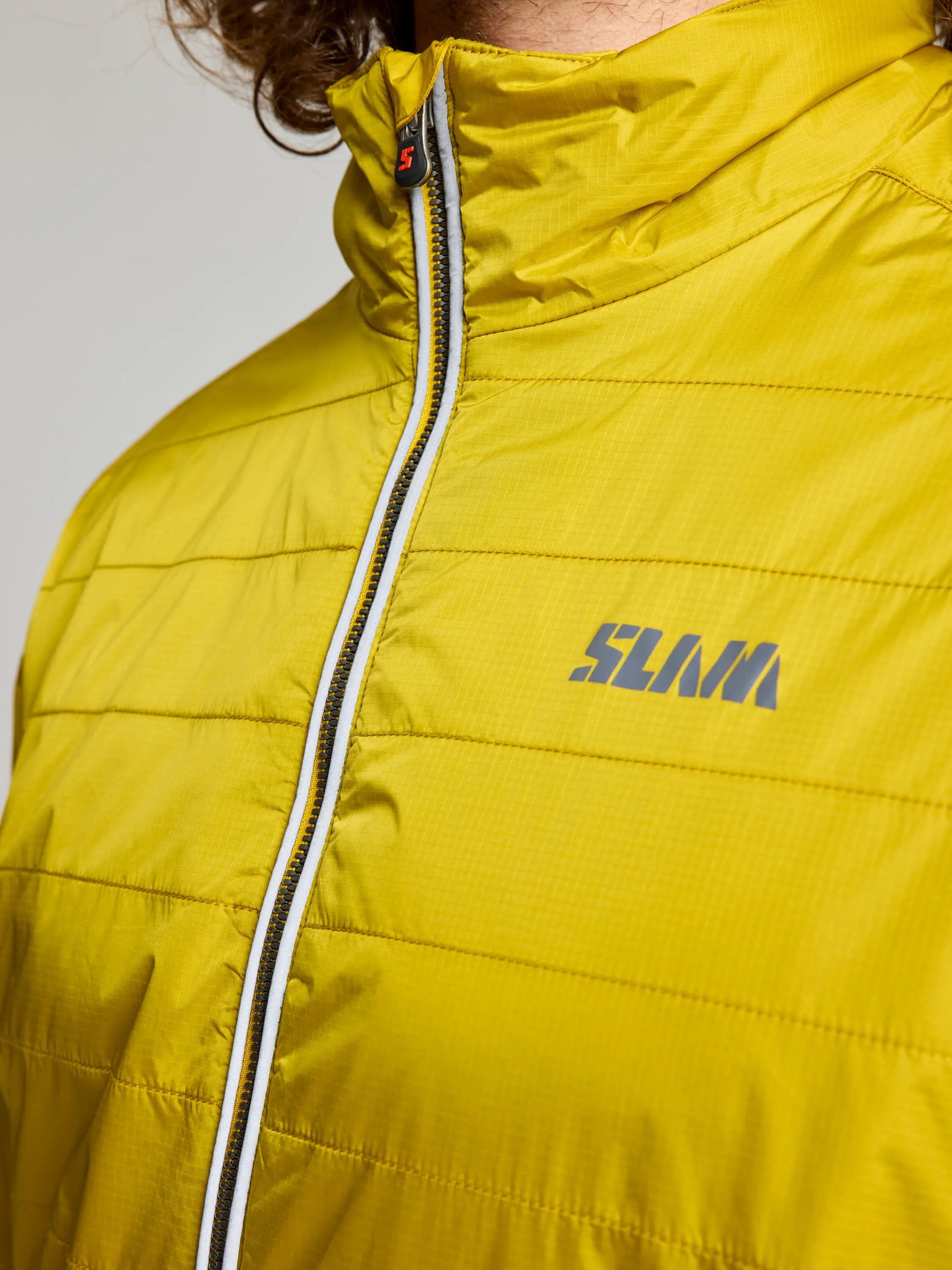 Close-up of a person wearing the bright yellow ACTIVE GRAPHENE SHORT JACKET CL by SLAM, with the logo clearly visible on the right chest area. The quilted texture of this recycled nylon ripstop jacket complements their curly hair peeking out.