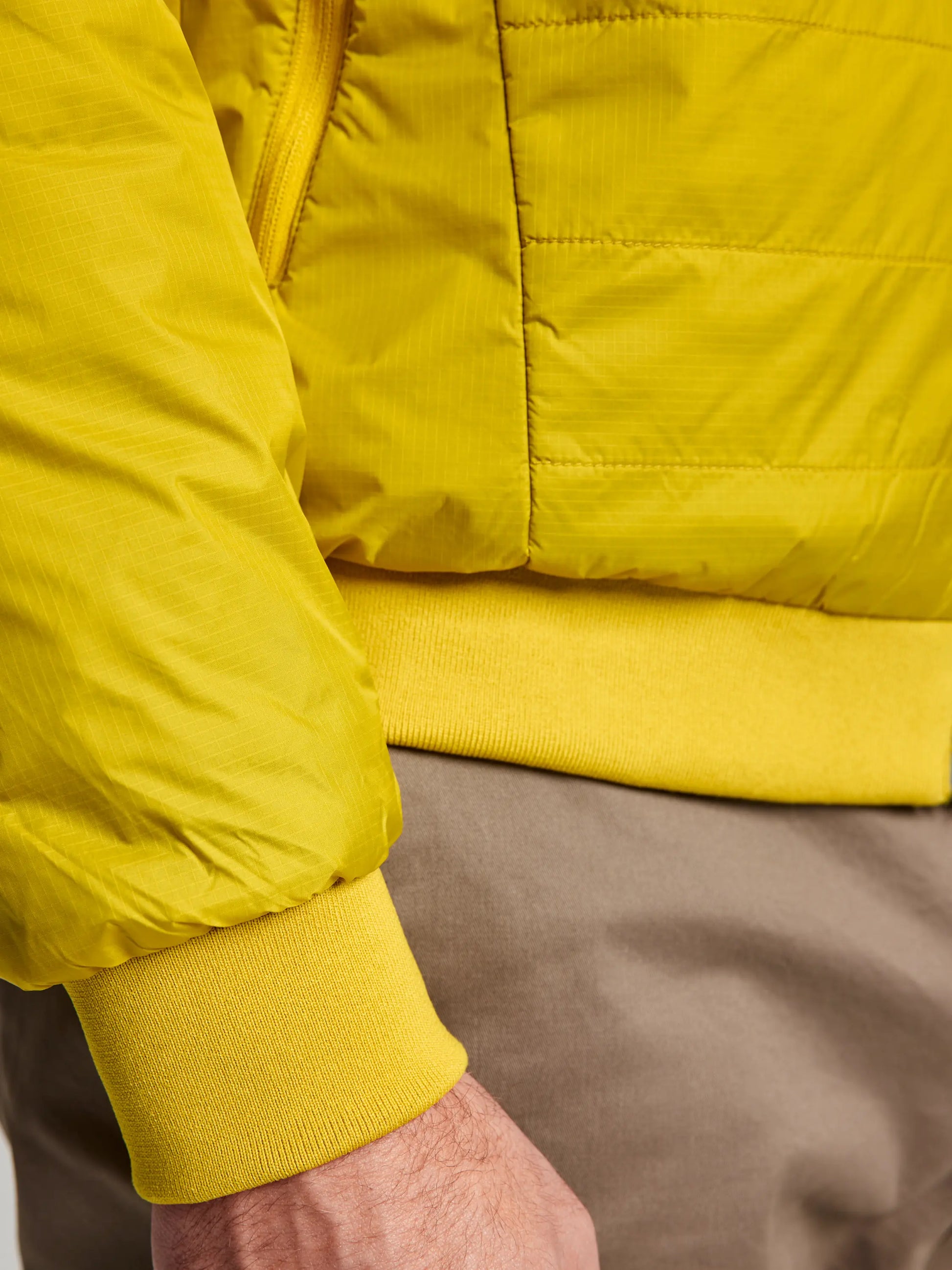 A close-up showcases a person wearing the ACTIVE GRAPHENE SHORT JACKET CL by SLAM, highlighting its vibrant yellow quilted design crafted from recycled nylon ripstop. The focus is on the sleeve and side pocket detail, complemented by light brown pants and a partially visible hand, adding a touch of sophistication to this sustainable style.