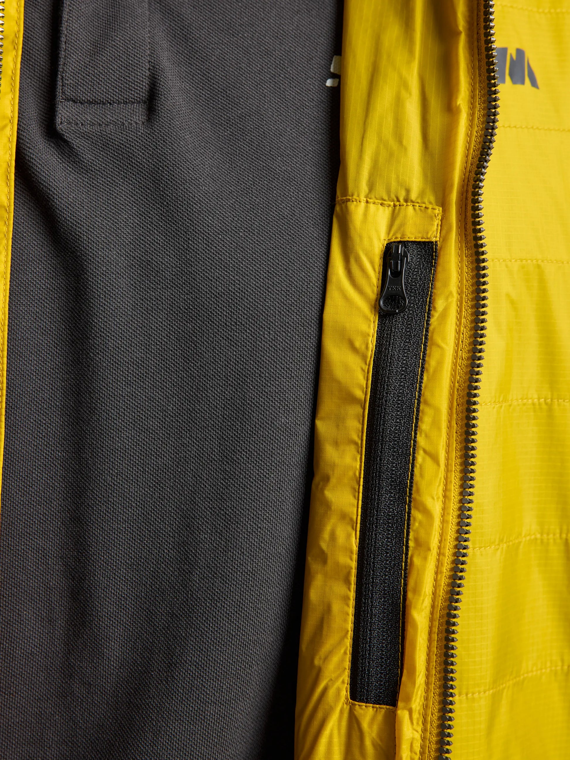 Close-up of a gray fabric shirt paired with the SLAM ACTIVE GRAPHENE SHORT JACKET CL in yellow, made from recycled nylon ripstop. The jacket's material has a quilted look due to polyester wadding, and a zipper pocket displays part of the logo.