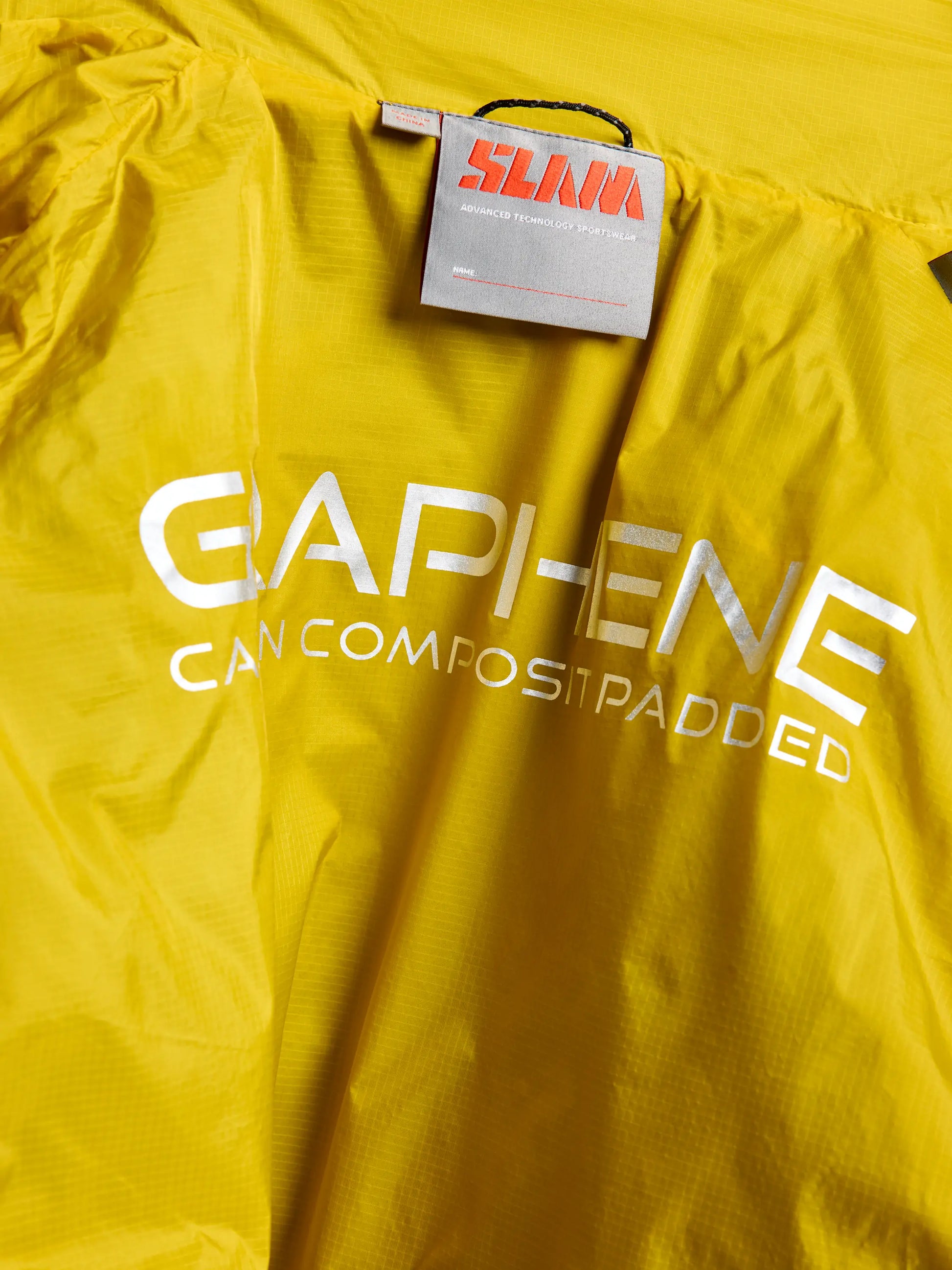 A close-up of a yellow ACTIVE GRAPHENE SHORT JACKET CL by SLAM, featuring a tag and a label with "GAPI-ENE CAN COMPOSIT PADDED" in white text. The fabric has a slightly shiny finish, crafted from recycled nylon ripstop, with a gray tag displaying red and white writing positioned above the label.