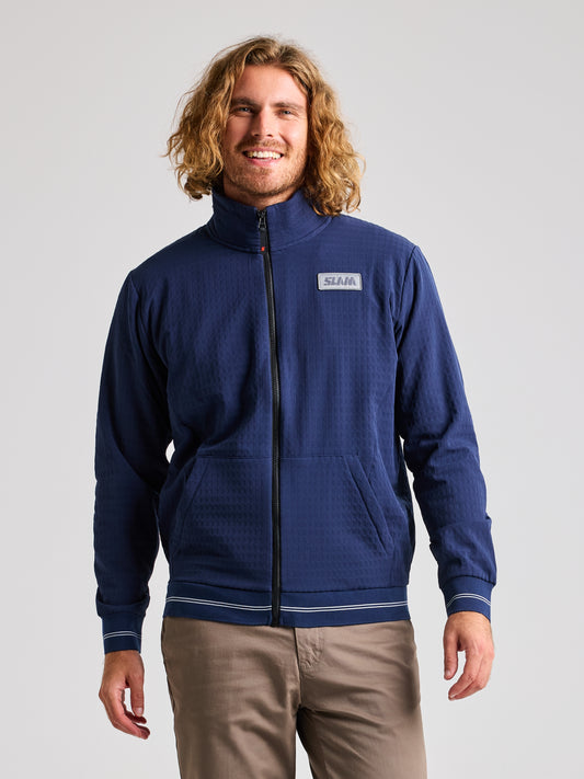 A person with long hair is sporting a fashionable SLAM MICKEY BEAR JACKET in Blue Shadow, featuring a high collar and a logo on the chest, expertly paired with beige pants. This nautical sportswear-inspired outfit pops against the plain, light-colored background.