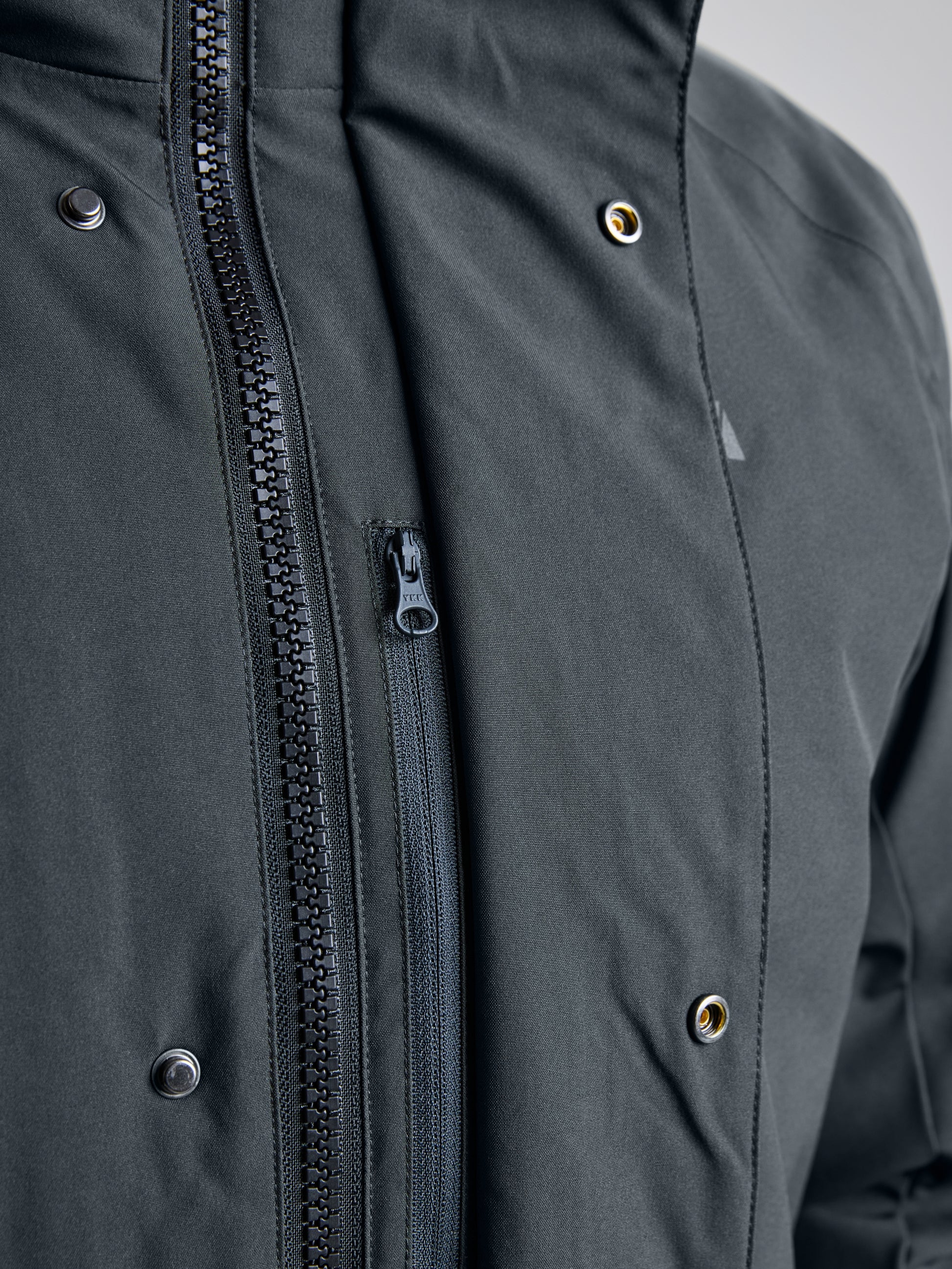 A close-up of the SLAM DECK COAT CL showcases a dark green waterproof jacket with a hood. This warm winter coat features a partially open vertical black zipper and two rows of metal snap buttons on each side. Crafted from recycled materials, the fabric appears smooth and weather-resistant.