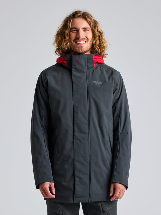 A person with long curly hair is wearing the DECK COAT CL by SLAM, a GRAPHITE waterproof jacket with a red hood, standing against a plain light gray background. Made from recycled fabrics, the jacket features a small logo on the chest. The person is smiling and has their hands by their sides.