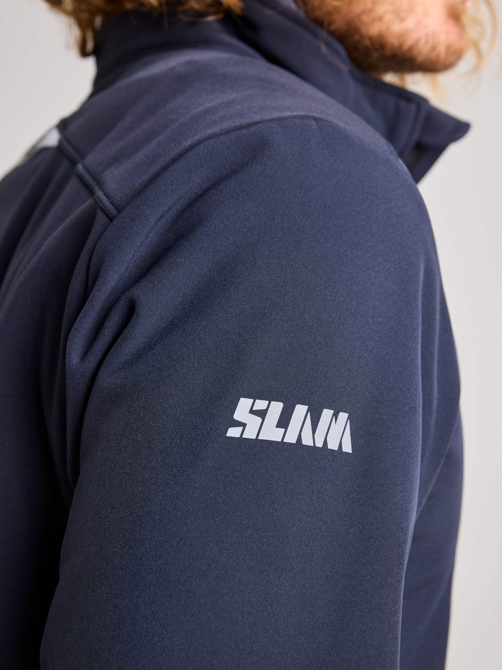 Close-up of a person wearing the ACTIVE WARM SOFTSHELL JACKET by SLAM in dark blue. "SLAM" is printed in white on the right sleeve, but the person's face is not visible.