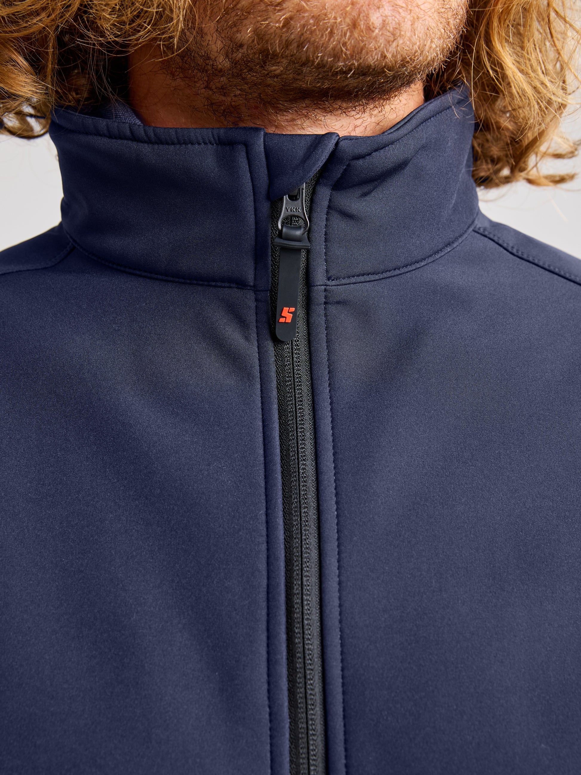 Close-up of a person wearing the SLAM ACTIVE WARM SOFTSHELL JACKET in navy blue, featuring a high collar. The waterproof fabric accentuates the zipper and material texture, while the person's chin and glimpse of long, wavy hair add character to the scene.