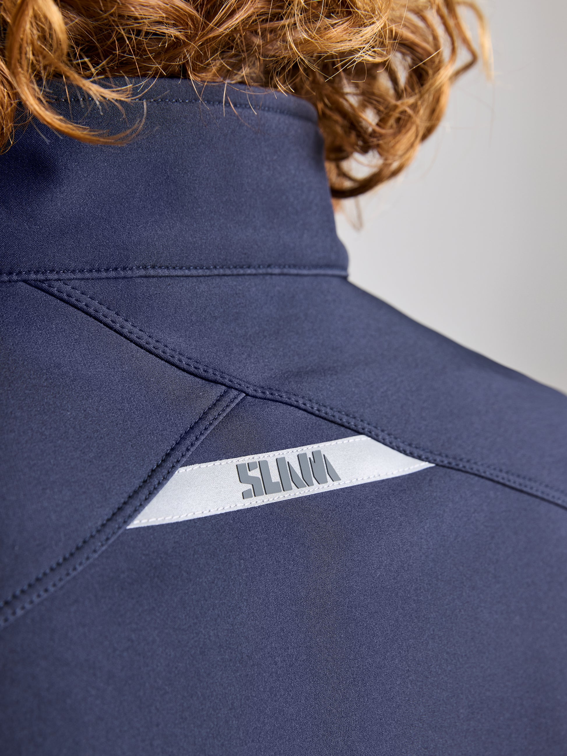 Close-up image showing the back of a person wearing the ACTIVE WARM SOFTSHELL JACKET from SLAM in navy blue, designed with a high collar. This breathable jacket incorporates a reflective strip and features "SILVA" printed upside down in silver letters, partially obscured by wavy hair.