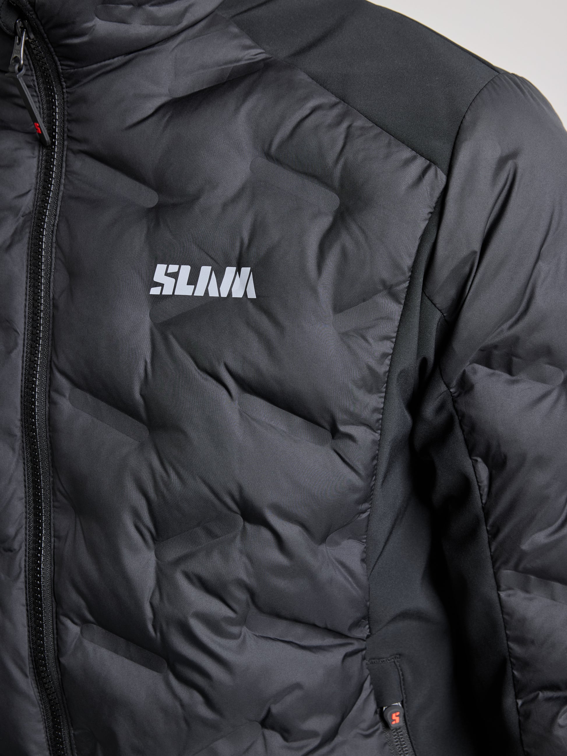 Close-up of the HYBRID SOFTSHELL JACKET in black, showcasing a visible zip and the brand "SLAM" in white lettering on the chest. This breathable jacket features a textured design with slightly shiny material, delivering both style and comfort.