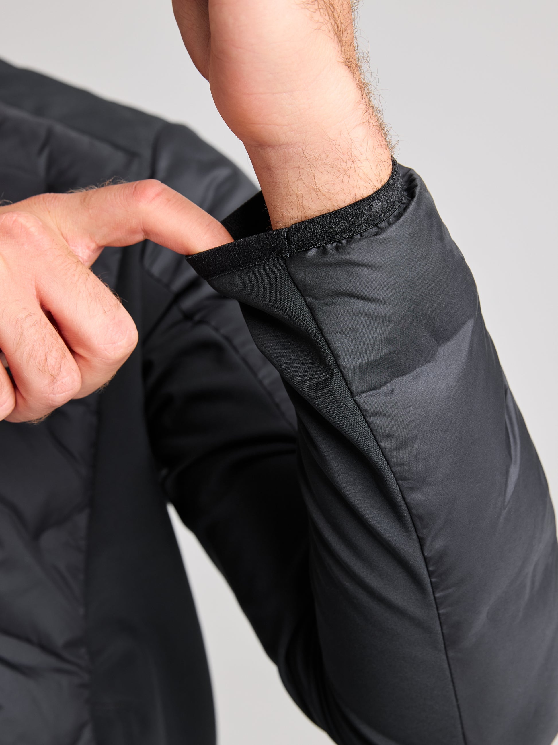 A person adjusts the cuff of a breathable HYBRID SOFTSHELL JACKET in black, using their right hand to pull on the left sleeve. Made by SLAM, the jacket material looks smooth and slightly shiny, ideal for any weather, against a neutral gray background.