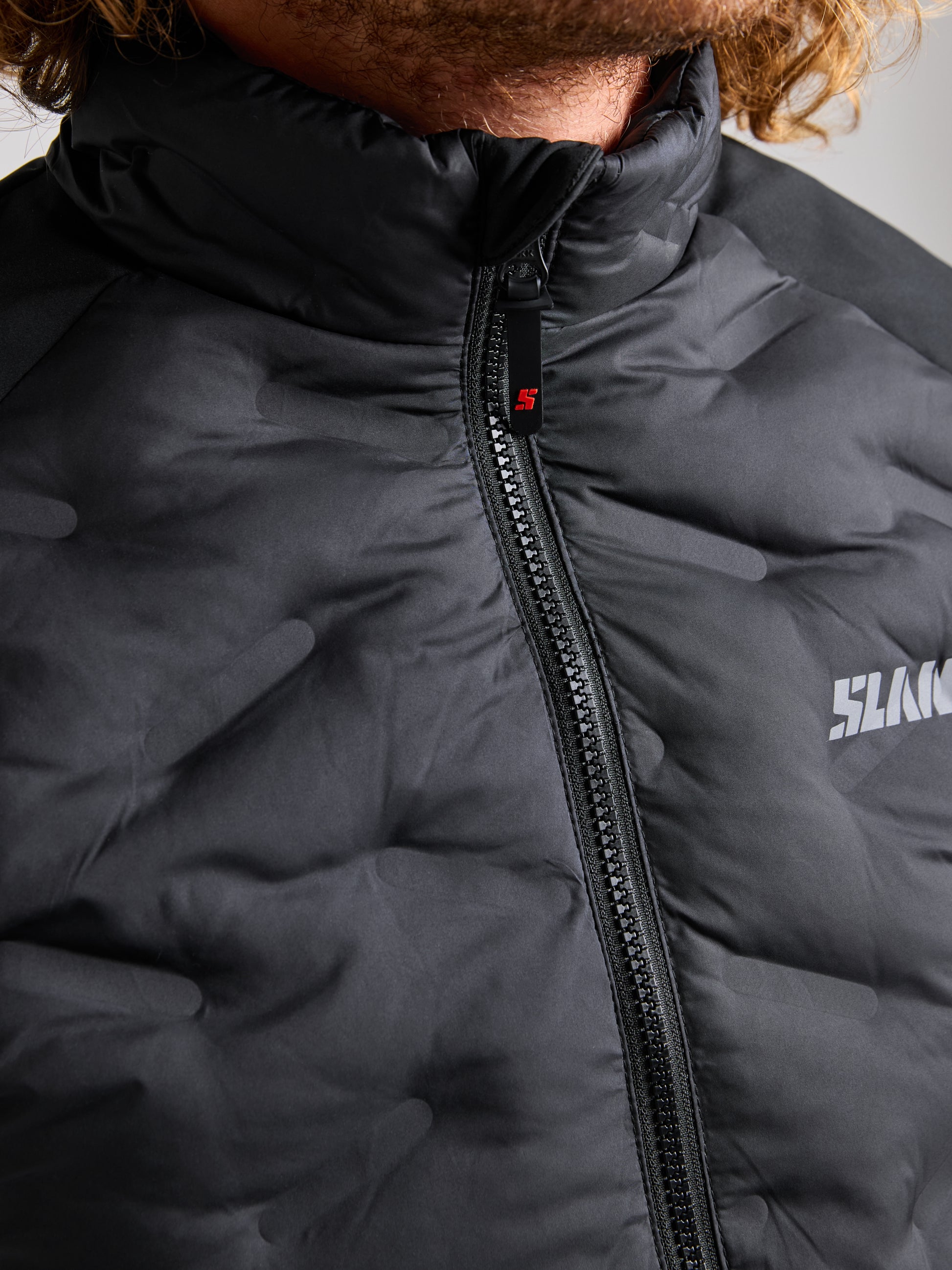 A close-up shot showcases an individual dressed in a black SLAM HYBRID SOFTSHELL JACKET, highlighting the visible zipper. This breathable and waterproof jacket sports a discreet logo on the upper right side. At the top edge of the image, part of the person's curly hair can be seen.