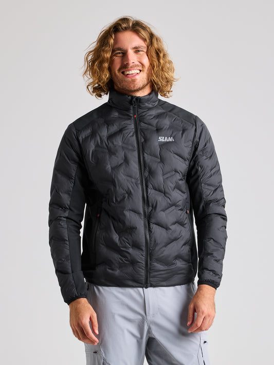 A person with long hair is wearing a BLACK INK quilted HYBRID SOFTSHELL JACKET by SLAM and light gray pants. They are standing against a plain background, smiling. This breathable jacket features the brand's logo on the left chest area.