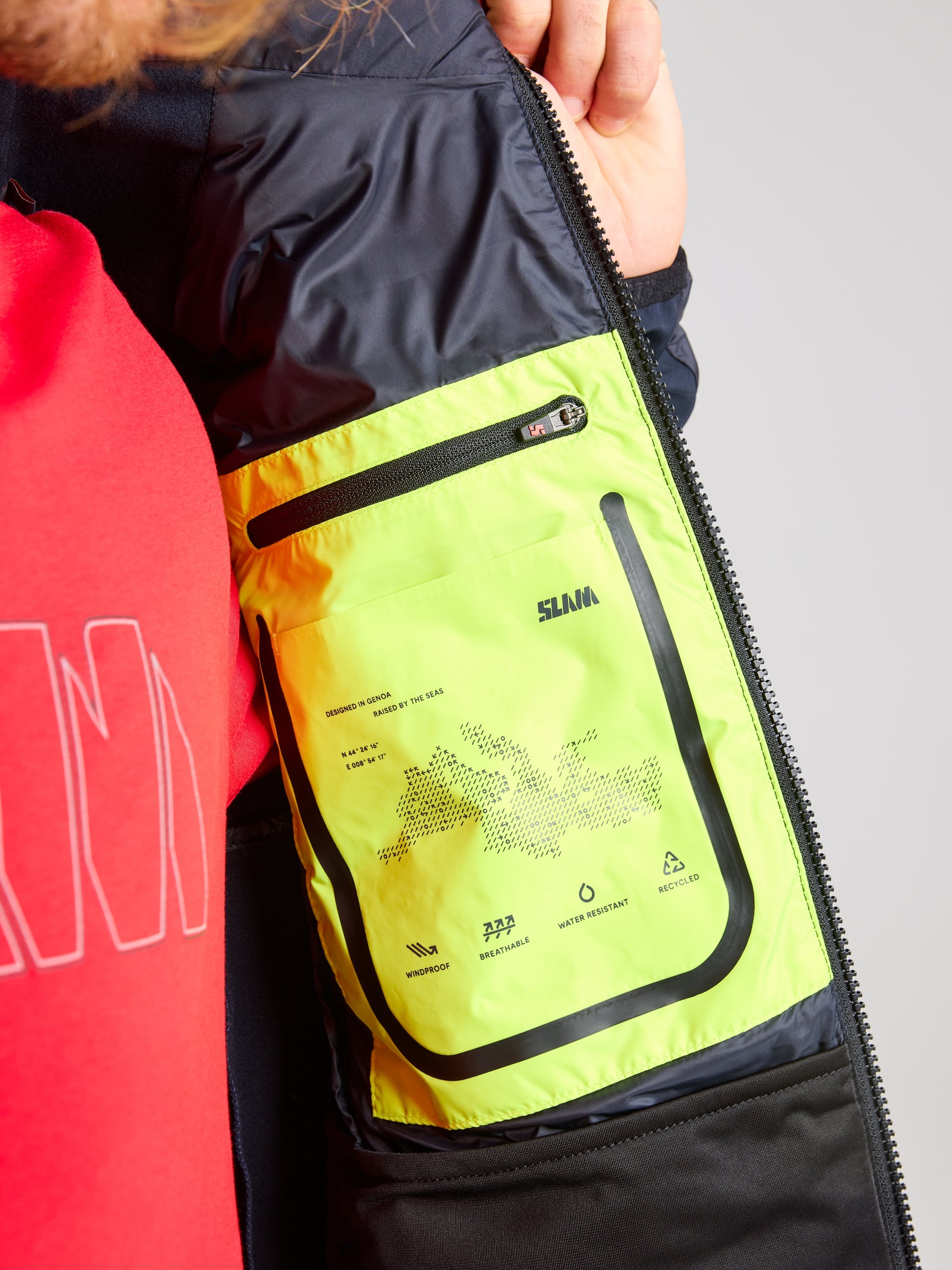 A person wearing a red shirt is opening a HYBRID SOFTSHELL JACKET, revealing an interior pocket that's brightly colored in neon yellow with black trim and zippers. The pocket features printed graphics and text, prominently displaying the brand name "SLAM." This breathable jacket seamlessly combines style with practicality.