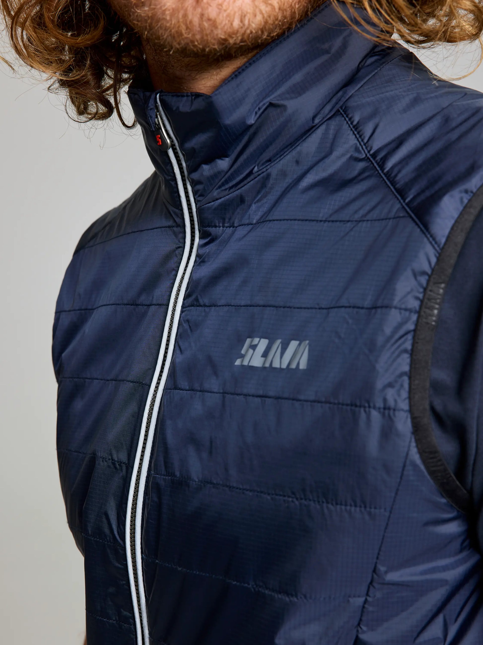 A person with curly hair is wearing a navy blue SLAM ACTIVE GRAPHENE RSTOP VEST, which features a white zipper and a subtle logo on the chest. The image highlights the upper chest and shoulder area of this men's gilet, emphasizing its sleek design crafted from recycled Nylon Ripstop.