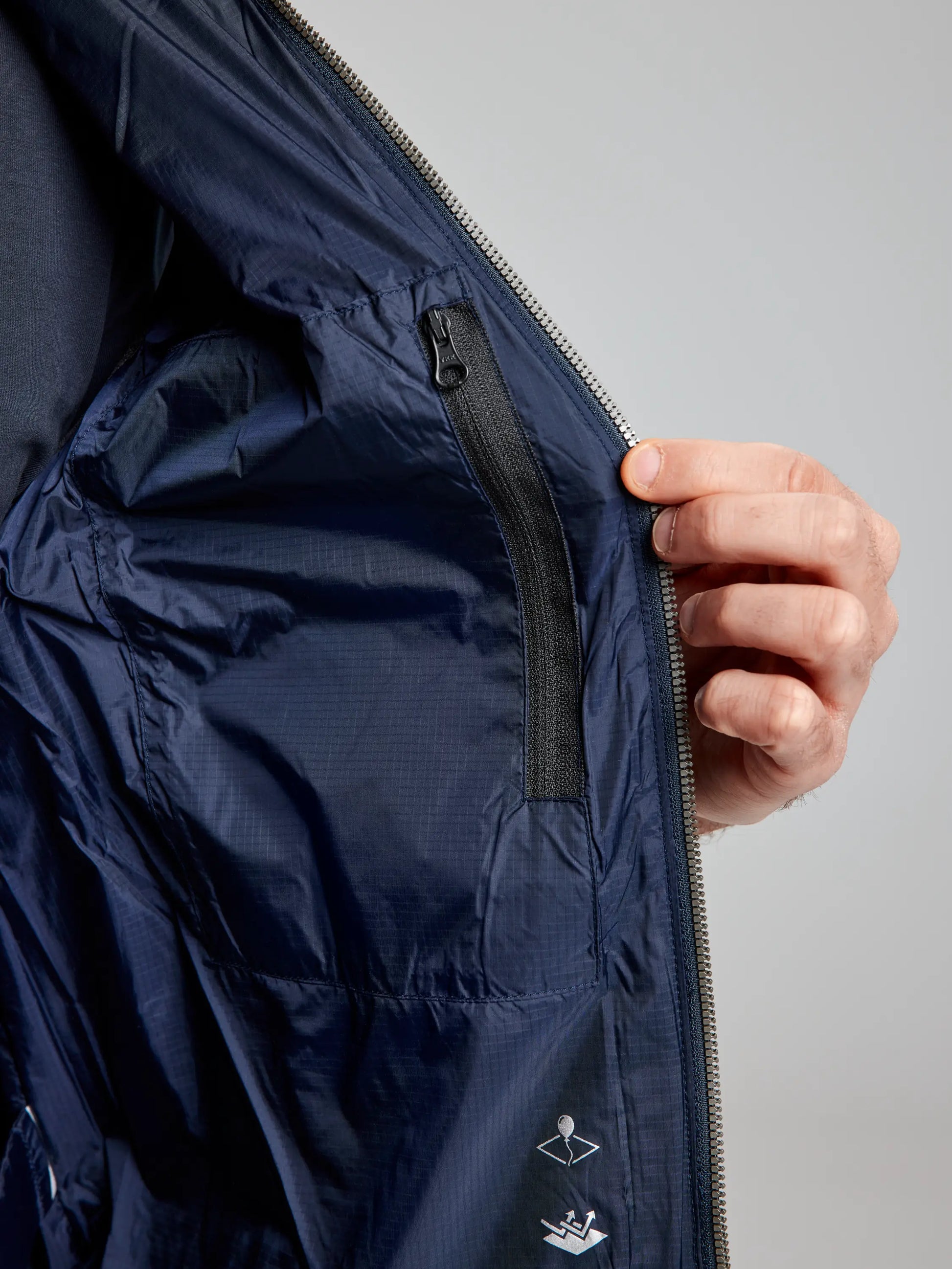 As someone opens a deep blue vest, they reveal the inner lining and a zippered pocket, designed with recycled Nylon Ripstop. This vest from SLAM has a subtle shiny texture and is embellished with small white symbols on the fabric.