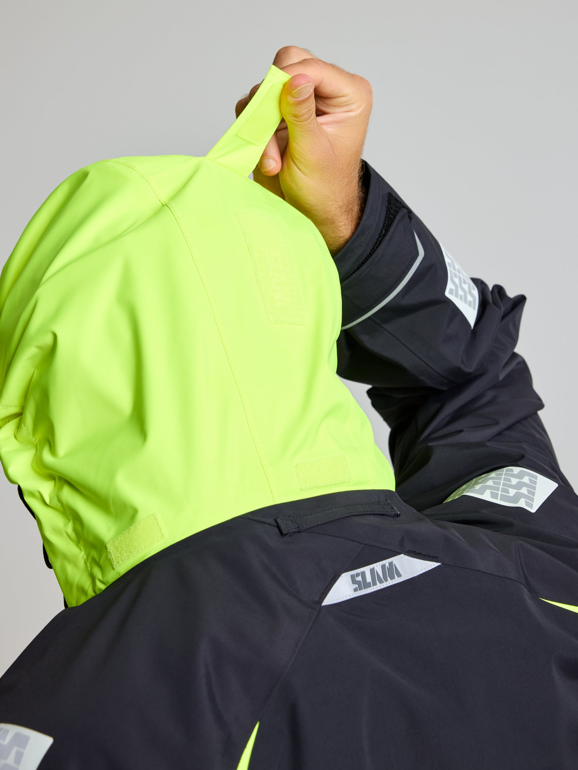 A person is adjusting the high-visibility hood of a neon yellow PRO OFFSHORE JACKET by SLAM. This sleek jacket is designed with gray patches and black sleeves, and prominently displays the brand name near the neckline. The plain, light-colored background highlights its stylish waterproof design.