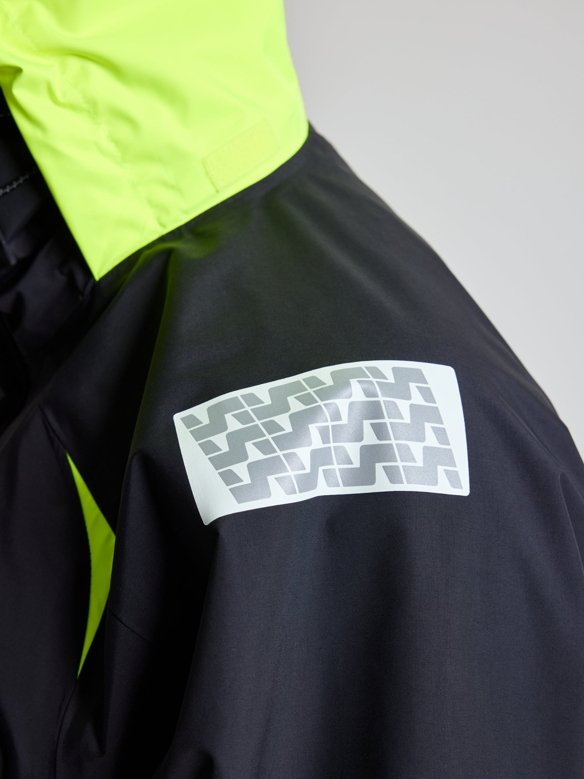 Close-up of the SLAM Pro Offshore Jacket, showcasing its neon yellow high-visibility hood and sleeve against a black body. A reflective patch featuring a geometric pattern adorns the shoulder, emphasizing the contrasting colors and design details of this waterproof garment.