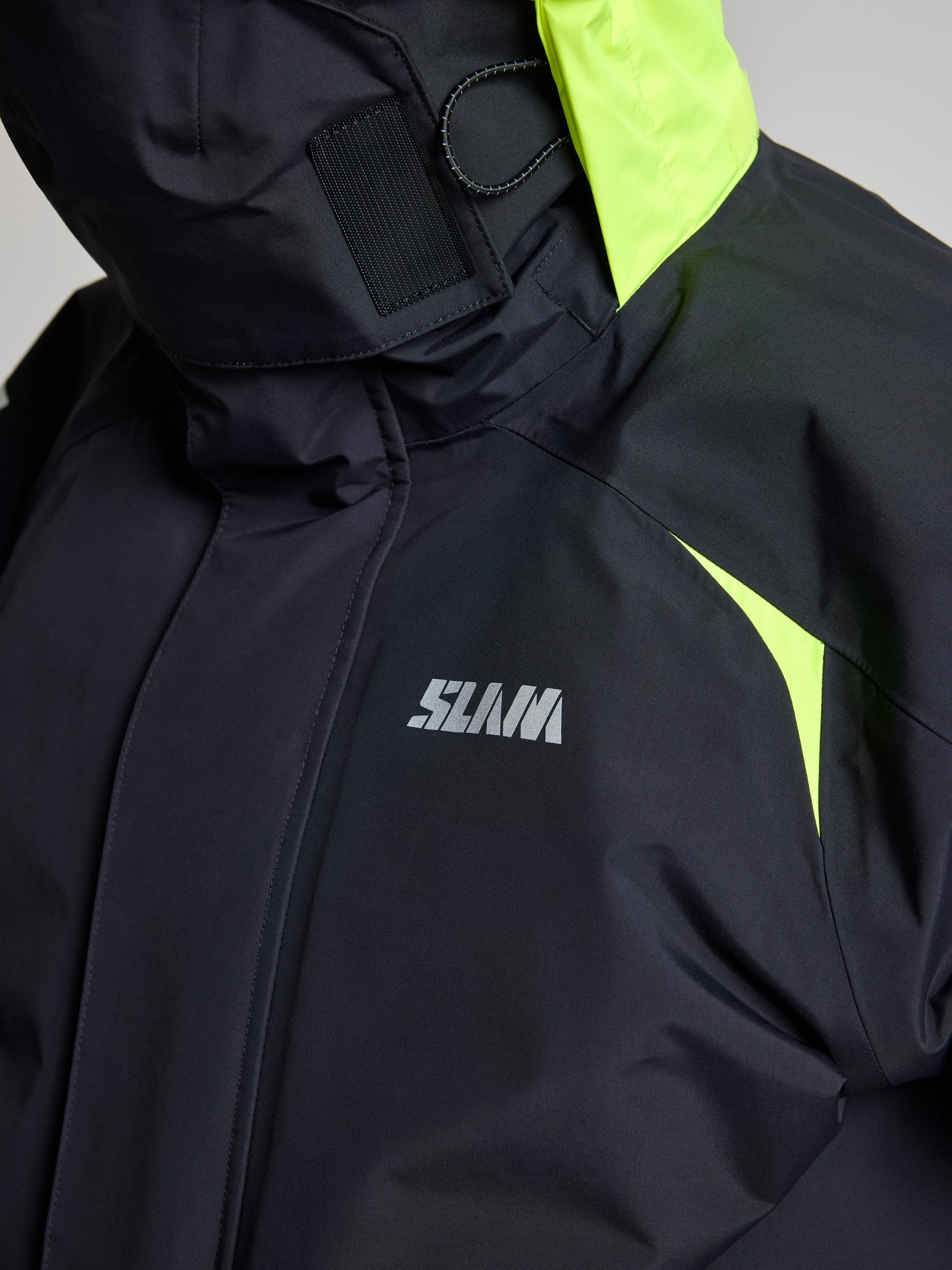 Close-up view of the SLAM PRO OFFSHORE JACKET in black, showcasing neon yellow accents on the shoulders and a high-visibility hood. The jacket displays a white logo on the chest, and the fabric seems to provide excellent waterproofing.