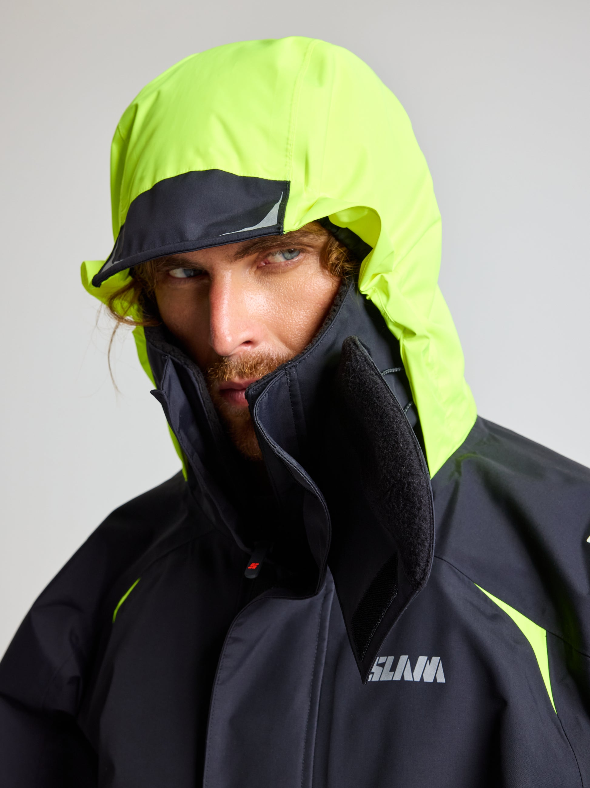 A person is wearing a neon yellow and black SLAM PRO OFFSHORE JACKET, complete with a high-visibility hood that's pulled up, partially obscuring their face. The jacket prominently displays the SLAM logo on the chest area against a plain, light-colored background.