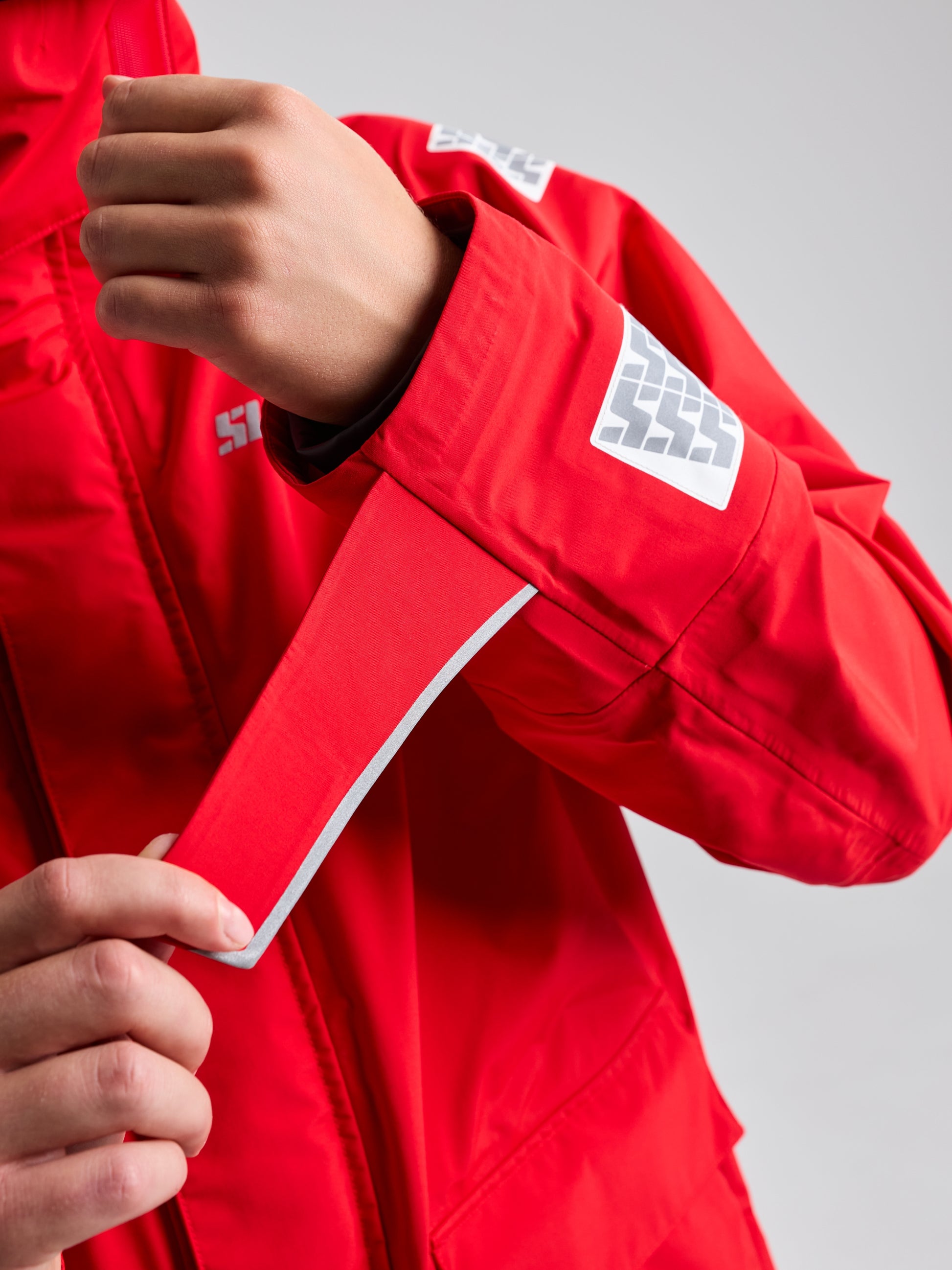 A person wearing a vibrant red SLAM PRO OFFSHORE JACKET with reflective patches adjusts the Velcro strap on the sleeve. The jacket, boasting a high-visibility hood and waterproofing, has a sleek and sporty design perfect for outdoor activities.