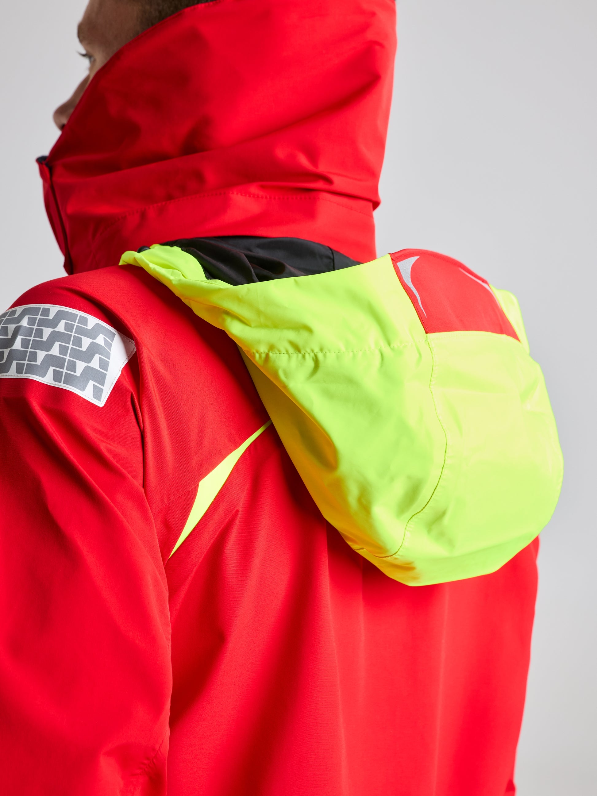 A person is seen from the side wearing SLAM's PRO OFFSHORE JACKET in a vibrant red, complemented by a high-visibility hood. The jacket is accented with black detailing on the shoulder and a grey patterned patch on the sleeve, highlighting its excellent waterproofing properties. The person is facing to the left.