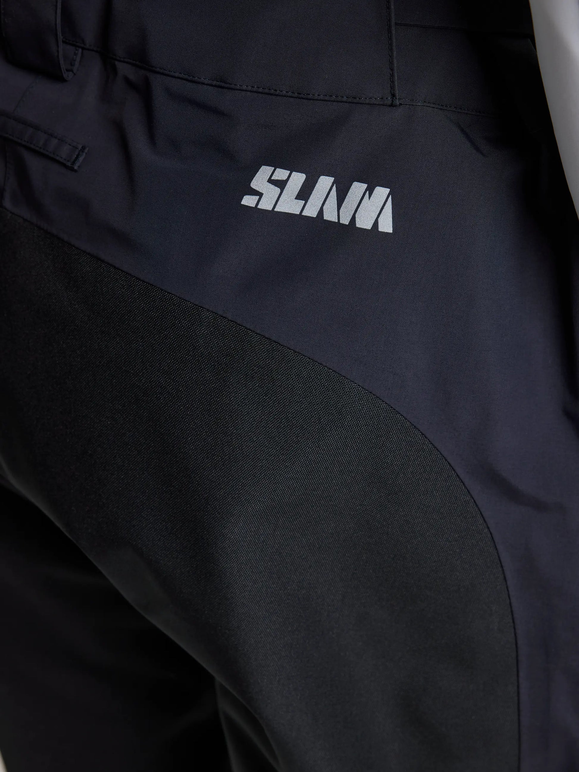 Close-up of the PRO OFFSHORE PANT by SLAM, showcasing dark tech nylon with a contrasting black panel. The word "SLAM" is printed in light-colored text near the waistband, providing protection against water, wind, and rain for performance in all weather conditions.