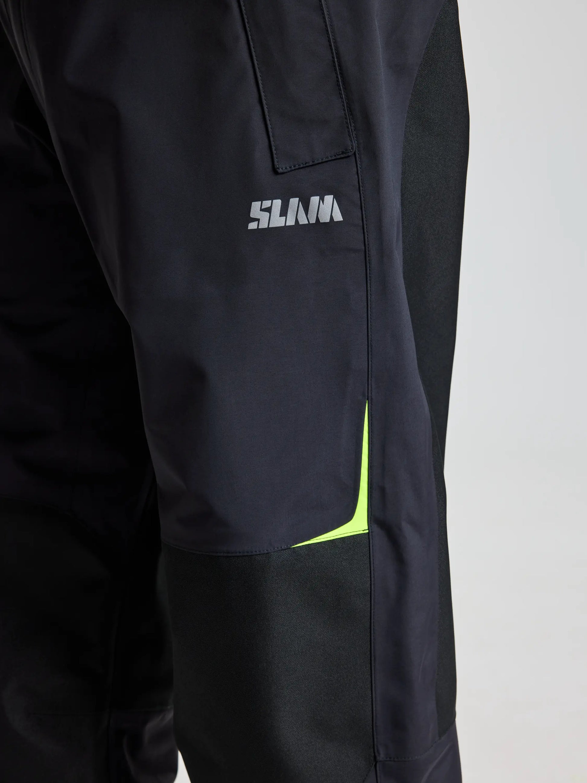 Close-up of a black fabric garment from the SLAM brand, featuring the "PRO OFFSHORE PANT" with gray letters. Accented with a yellow detail and visible stitching on the right side, this tech nylon pant provides protection against water, wind, and rain, indicating it is suitable for activewear or outerwear.