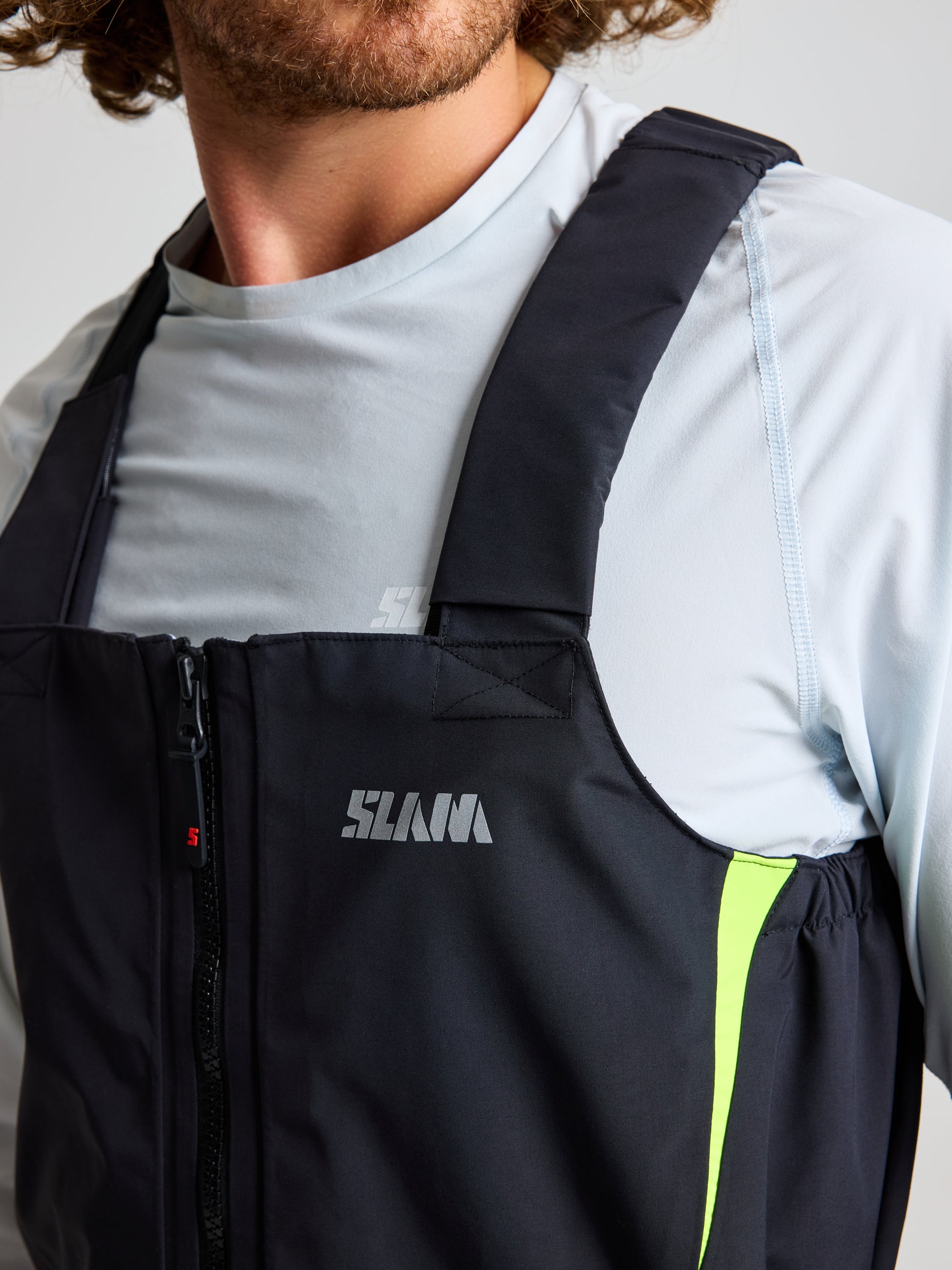 A person wearing a black and gray sleeveless jacket with a zipper, adorned with a logo on the front, layers it over a light gray long-sleeve shirt. The ensemble is completed with unisex PRO OFFSHORE BIBS from SLAM, which match the jacket's style and are distinguished by a bright green stripe along the side.