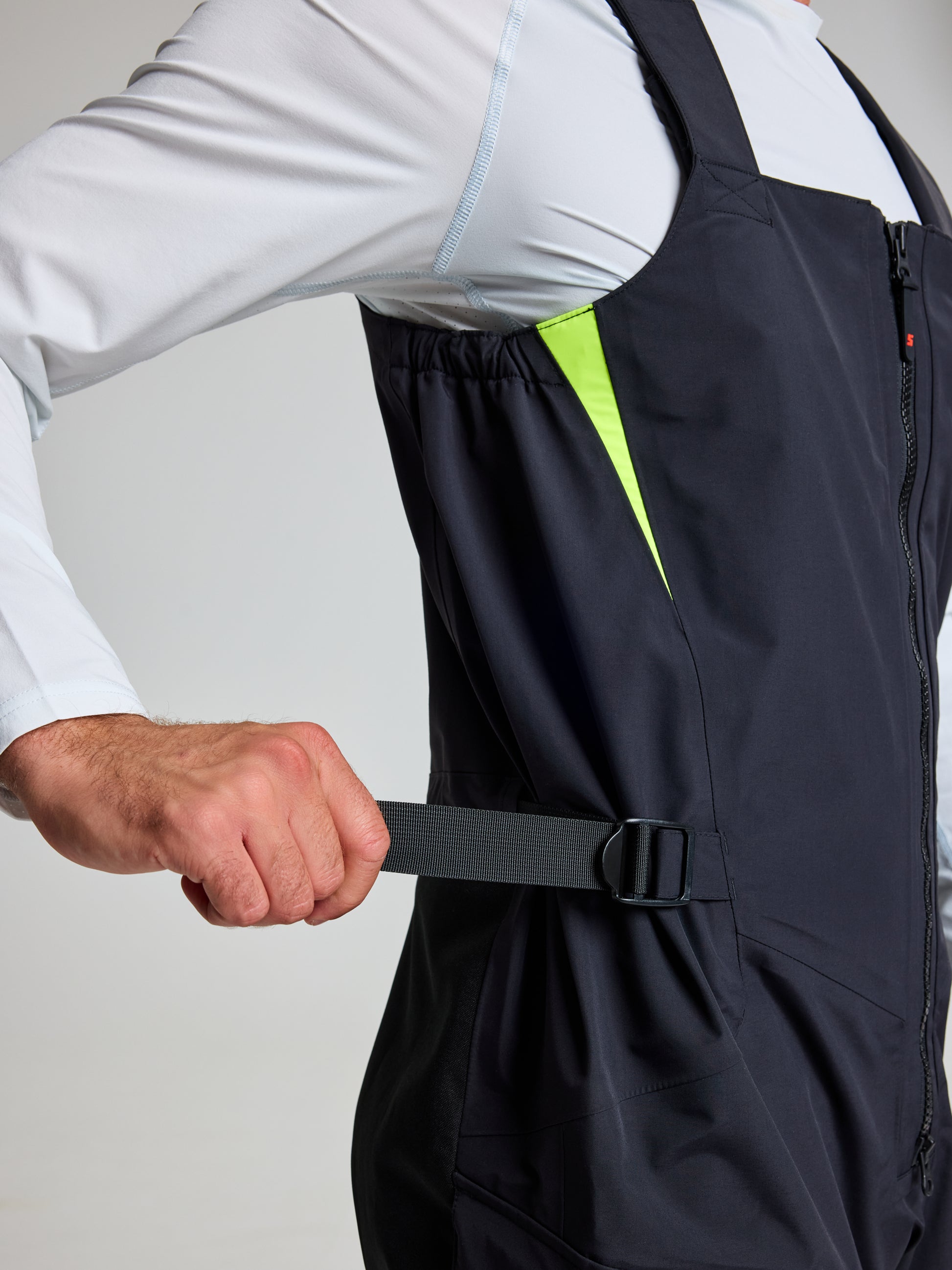 A person wearing the SLAM PRO OFFSHORE BIBS, resembling unisex overalls with a black zip-front design, adjusts a strap on the side. The bibs feature a neon yellow lining visible under the arm, while a white long-sleeve shirt peeks out underneath.