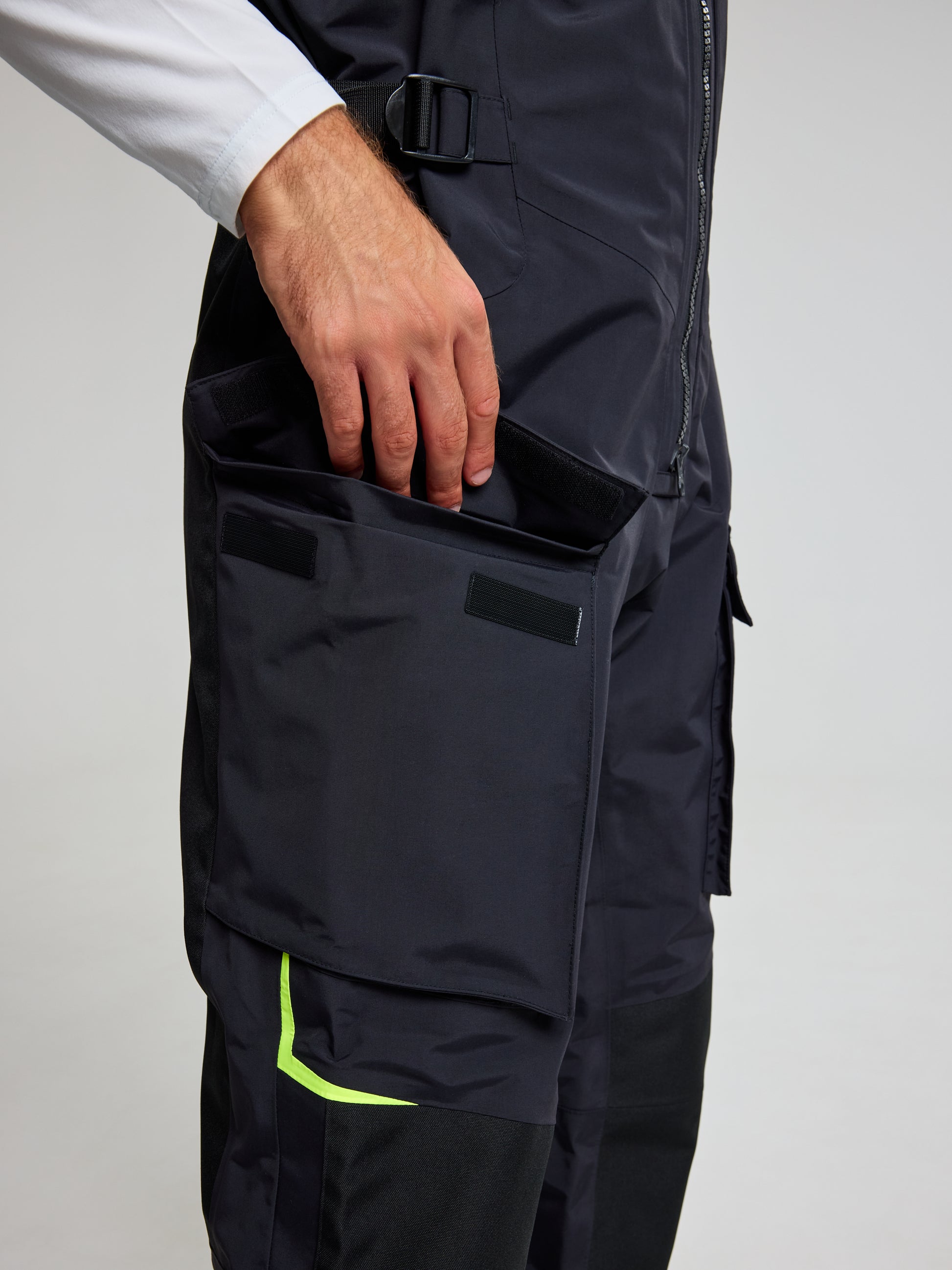 A person wearing the PRO OFFSHORE BIBS by SLAM, which resembles unisex overalls, is adjusting a large side pocket with a Velcro closure. The bibs feature a lime green accent on the sleeve, and the image provides a close-up view of the pocket and part of the arm.