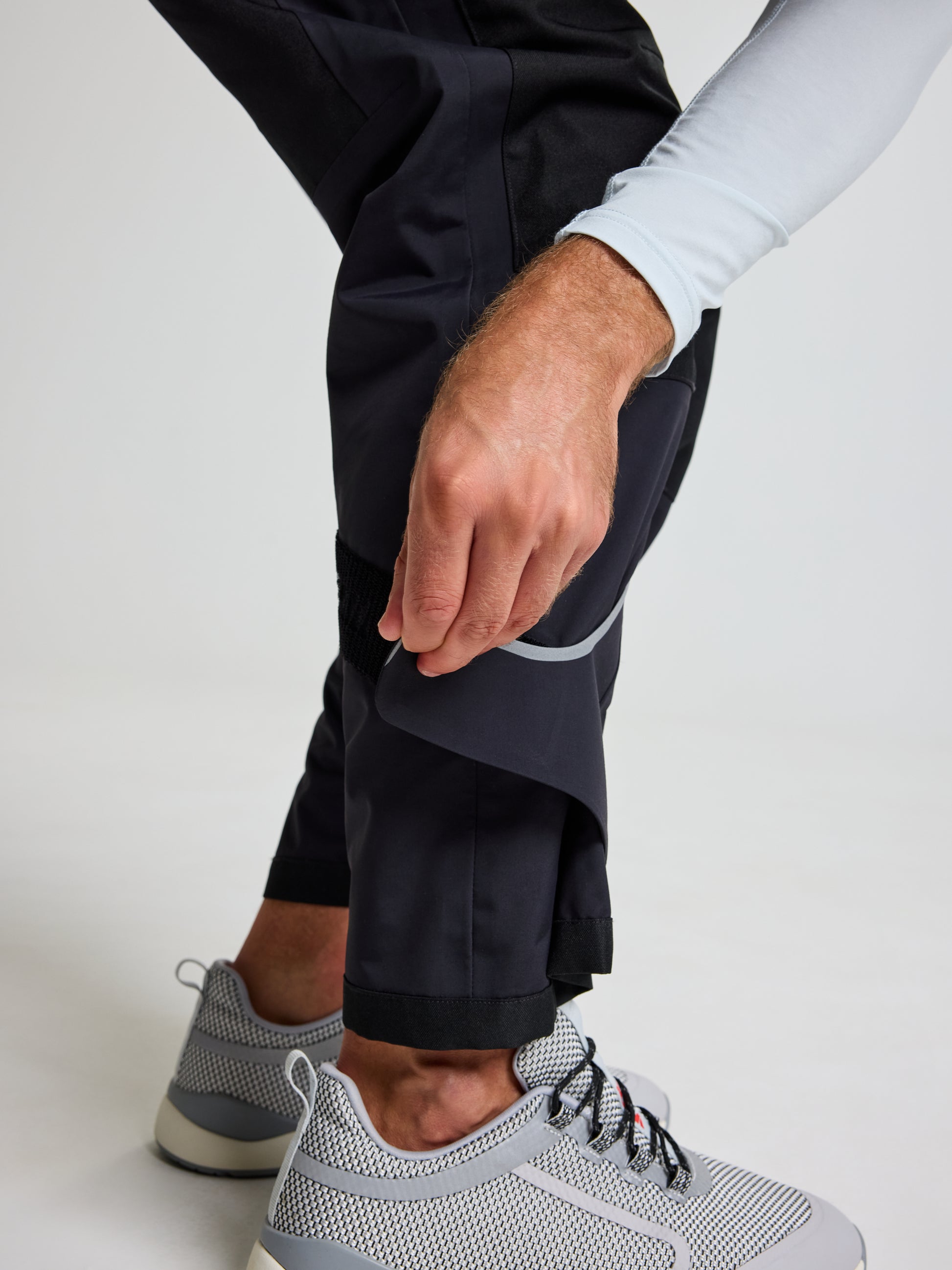 A person wearing SLAM's PRO OFFSHORE BIBS in black and light gray sneakers with a textured pattern and black laces adjusts the hem of their unisex overalls. The individual is dressed in a light-colored long-sleeve shirt, set against a plain and neutral background.