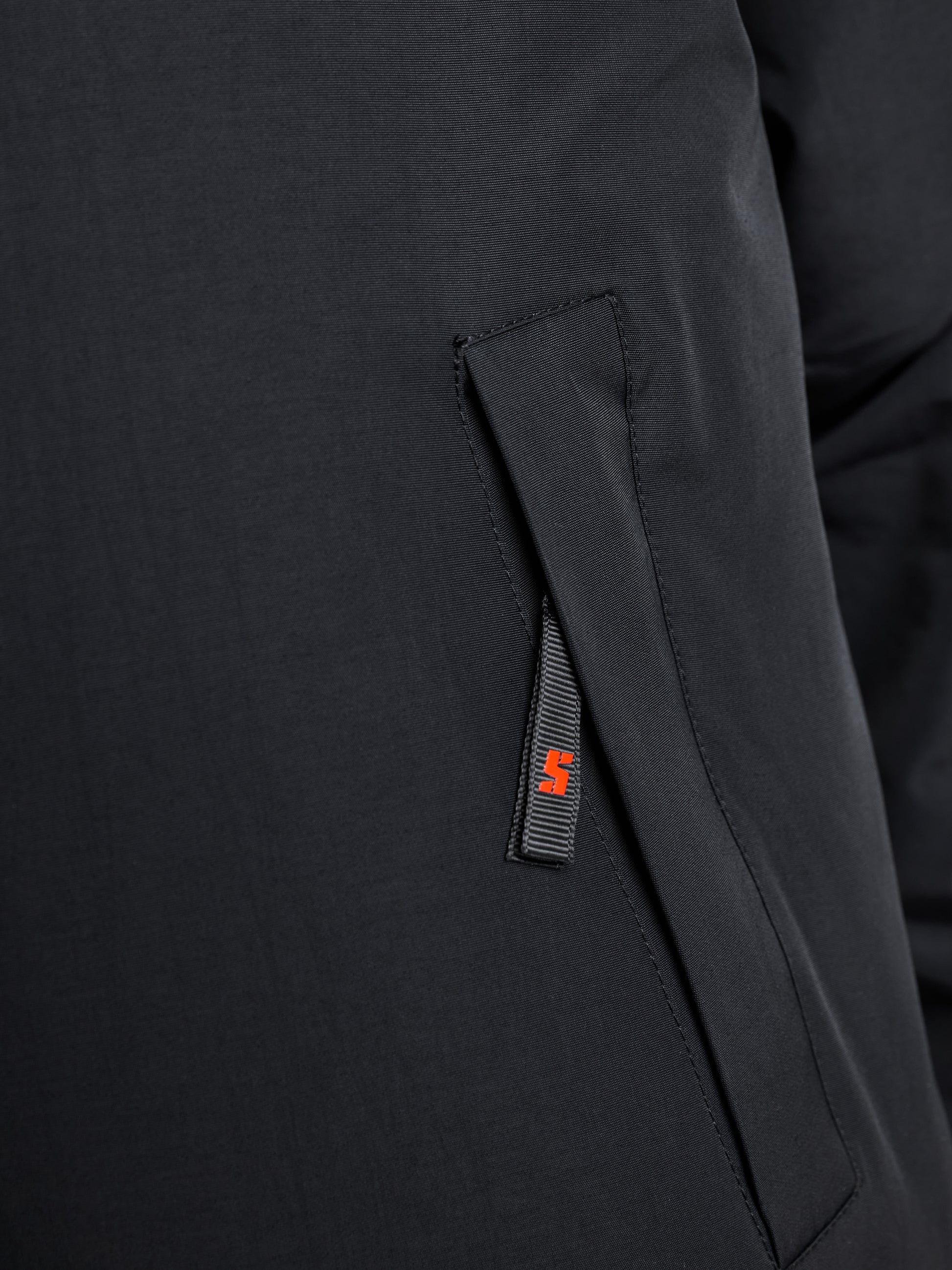 The close-up image showcases the JUNIOR SAILING WARM JACKET by SLAM, crafted from sleek, water-resistant Taslon 228T nylon. It includes a side pocket with a vertical flap, highlighted by a small red tag featuring an abstract design attached to the seam. The fabric has a smooth and slightly shiny appearance.