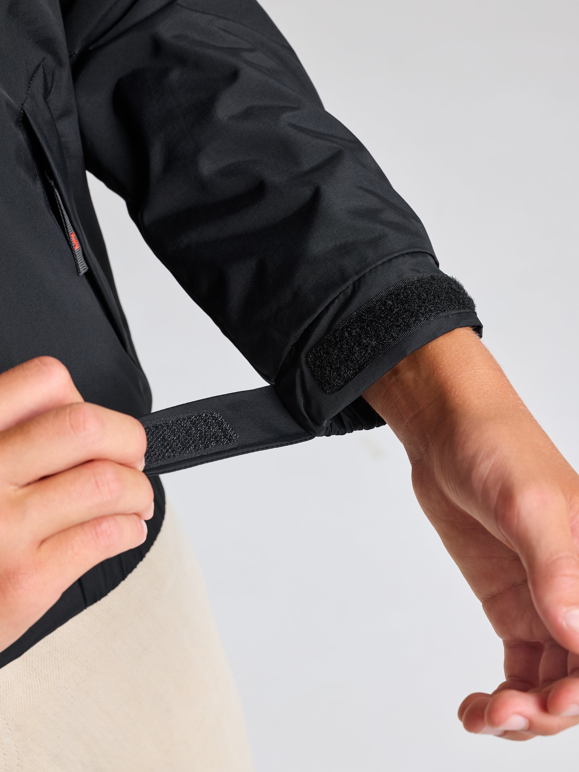 A person is adjusting the Velcro strap on the cuff of a black JUNIOR SAILING WARM JACKET from SLAM, crafted with water-resistant Taslon 228T nylon. They are wearing light-colored pants against a soft gray background.