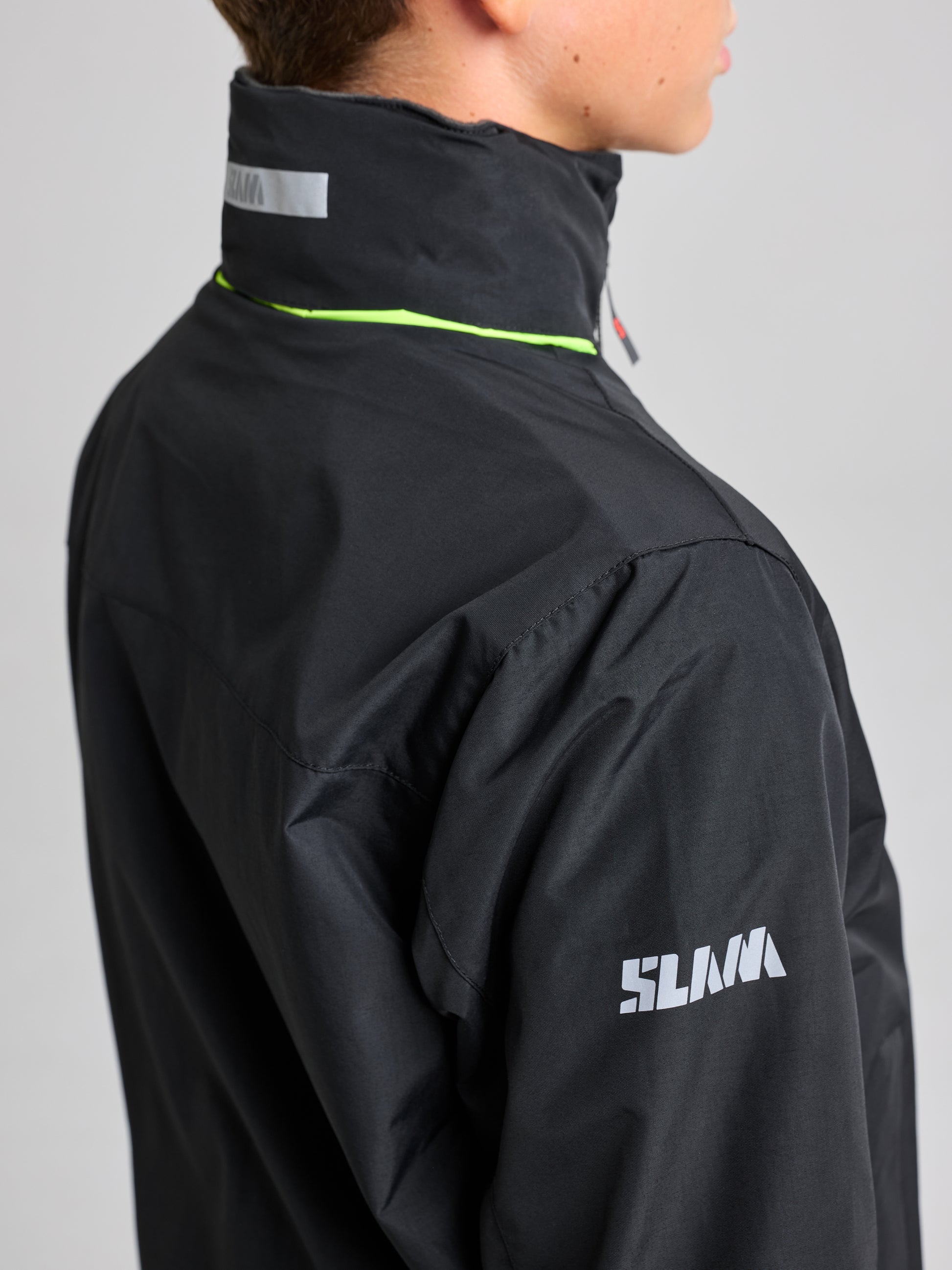 A person is depicted wearing the Junior Sailing Warm Jacket in water-resistant black, made from durable Taslon 228T nylon. Seen from the back and side, the jacket showcases "SLAM" in white lettering on its high collar and sleeve, highlighted with a lime green stripe. The plain gray background enhances its sleek design.