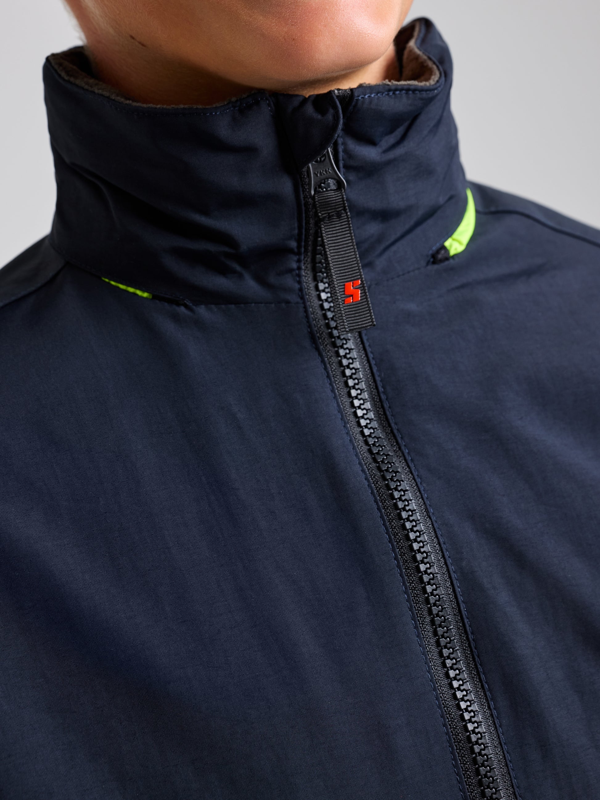 Close-up view of a person wearing the JUNIOR SAILING WARM JACKET by SLAM, made from Taslon 228T nylon. The dark zip-up jacket includes a high collar and water-resistant fabric, highlighted with a small orange zipper detail and a neon green accent near the collar. The background is a neutral gray.