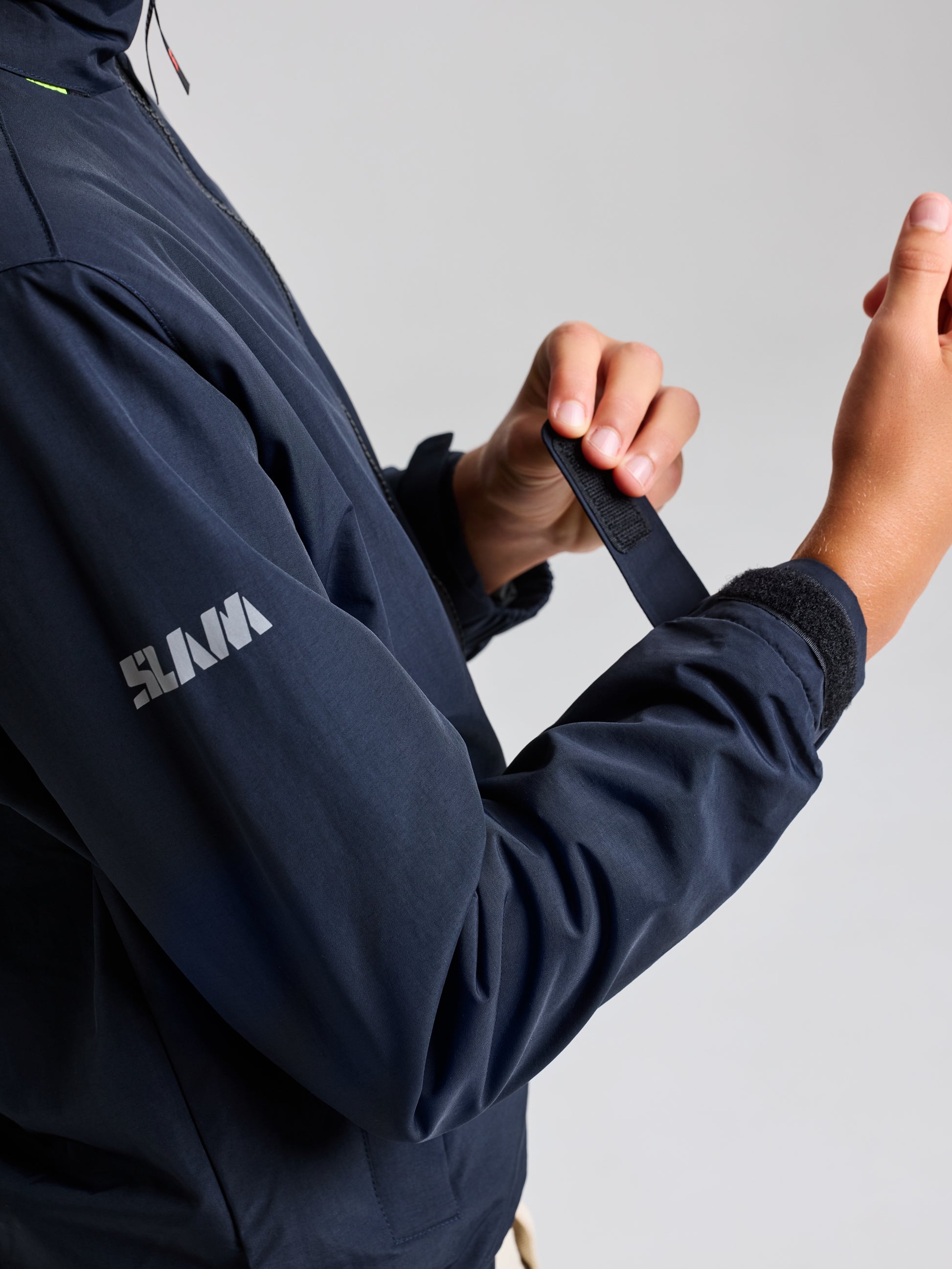 A person adjusts the Velcro strap on the cuff of a navy-blue JUNIOR SAILING WARM JACKET, made from Taslon 228T nylon with water-resistant properties. The word "SLAM" is printed on the arm, set against a plain and neutral backdrop.