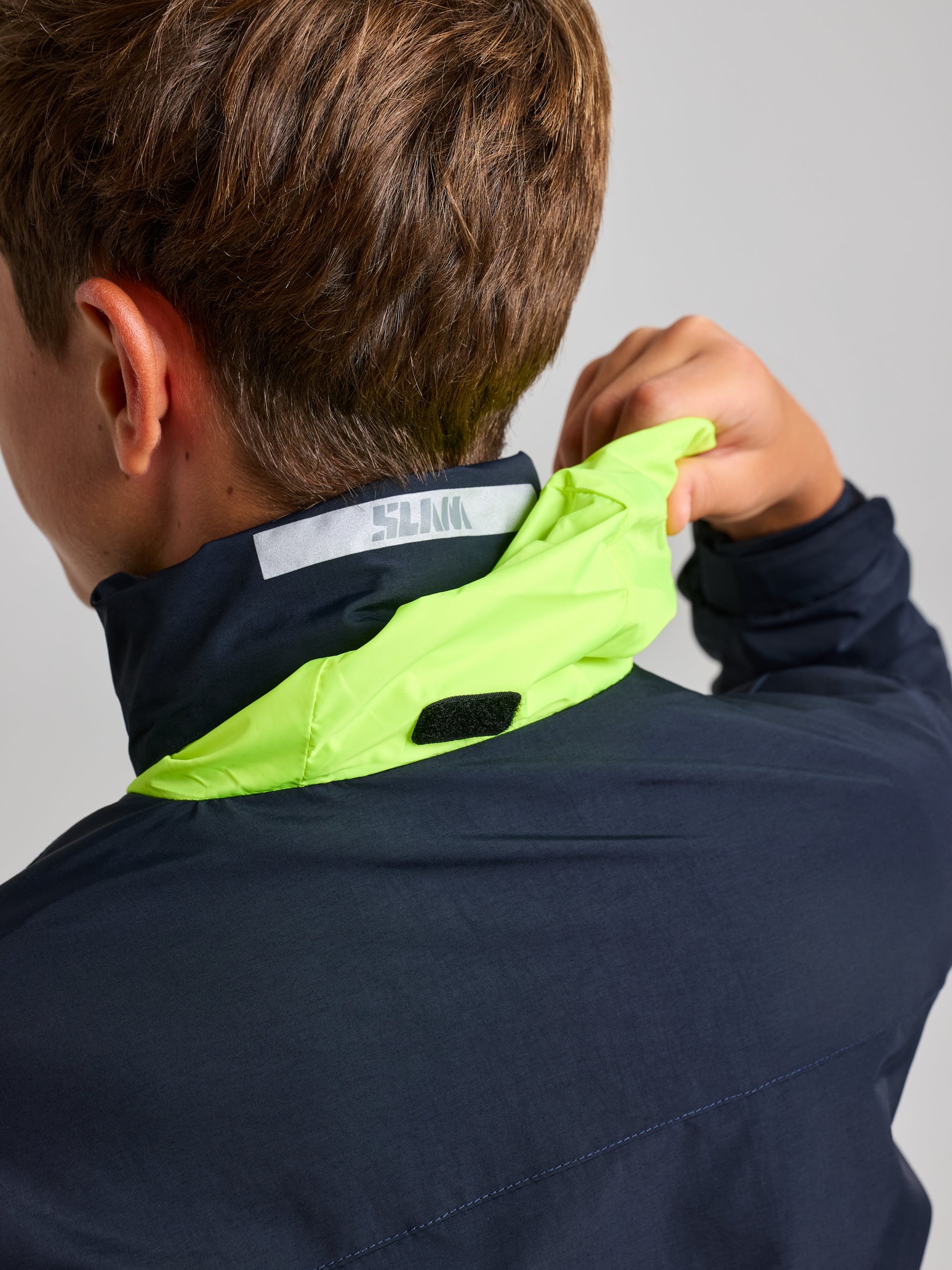 A person is seen from the back wearing a SLAM JUNIOR SAILING WARM JACKET, which has an adjustable neon green hood. Made from water-resistant Taslon 228T nylon, they're holding the hood with one hand while reflective text adorns the back of the collar.